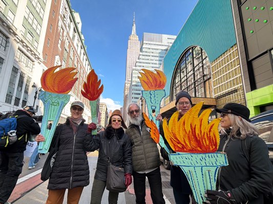 Five individuals are holding paper Statue of Liberty torches with stylized flames on a city street. In the background, the Empire State Building is visible. The people are standing on an urban road. The city environment includes buildings with green accents and windows. People wear coats and hats, appropriate for the weather, and gloves. The attire appears casual.