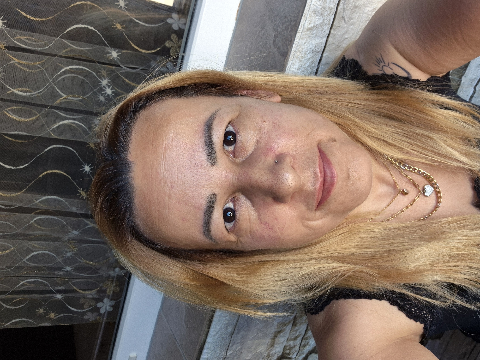 An outdoor, close-up selfie of a woman with blonde hair and fair skin. She is wearing a black lace top and several gold necklaces, one with a heart-shaped pendant. She has a nose ring and a small tattoo of a sun on her right shoulder. The background consists of a brick wall and sheer curtains with a floral pattern. The lighting suggests it is daytime.