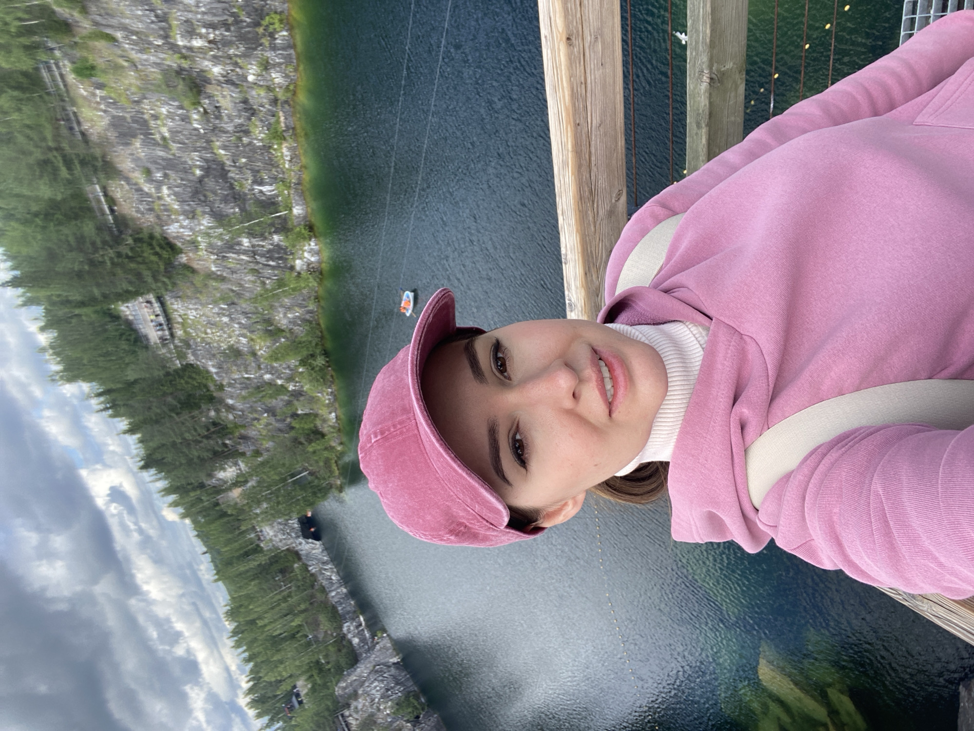 In the city of Ruskeala, Russia, a young woman with brown hair tied in a ponytail is taking a selfie on a wooden bridge overlooking a picturesque landscape. She is wearing a pink baseball cap, a white turtleneck, and a pink hooded sweatshirt. A beige backpack strap is visible over her shoulder.
The background reveals a stunning view of the Ruskeala Mountain Park, known for its marble canyon. A dark, serene lake fills the canyon, reflecting the surrounding lush green pine trees and the dramatic, rocky cliffs. The cliffs are characterized by their grey, textured surface, with some sections showing exposed white marble.
In the distance, a small boat with two people can be seen on the lake. A tunnel entrance is visible carved into the cliff face on the left side of the frame. Elevated wooden walkways with railings are constructed along the edges of the canyon, allowing visitors to traverse the area. The sky above is partly cloudy, with a mix of grey and white clouds against a pale blue backdrop. The overall scene captures the natural beauty and recreational aspects of the Ruskeala Mountain Park.