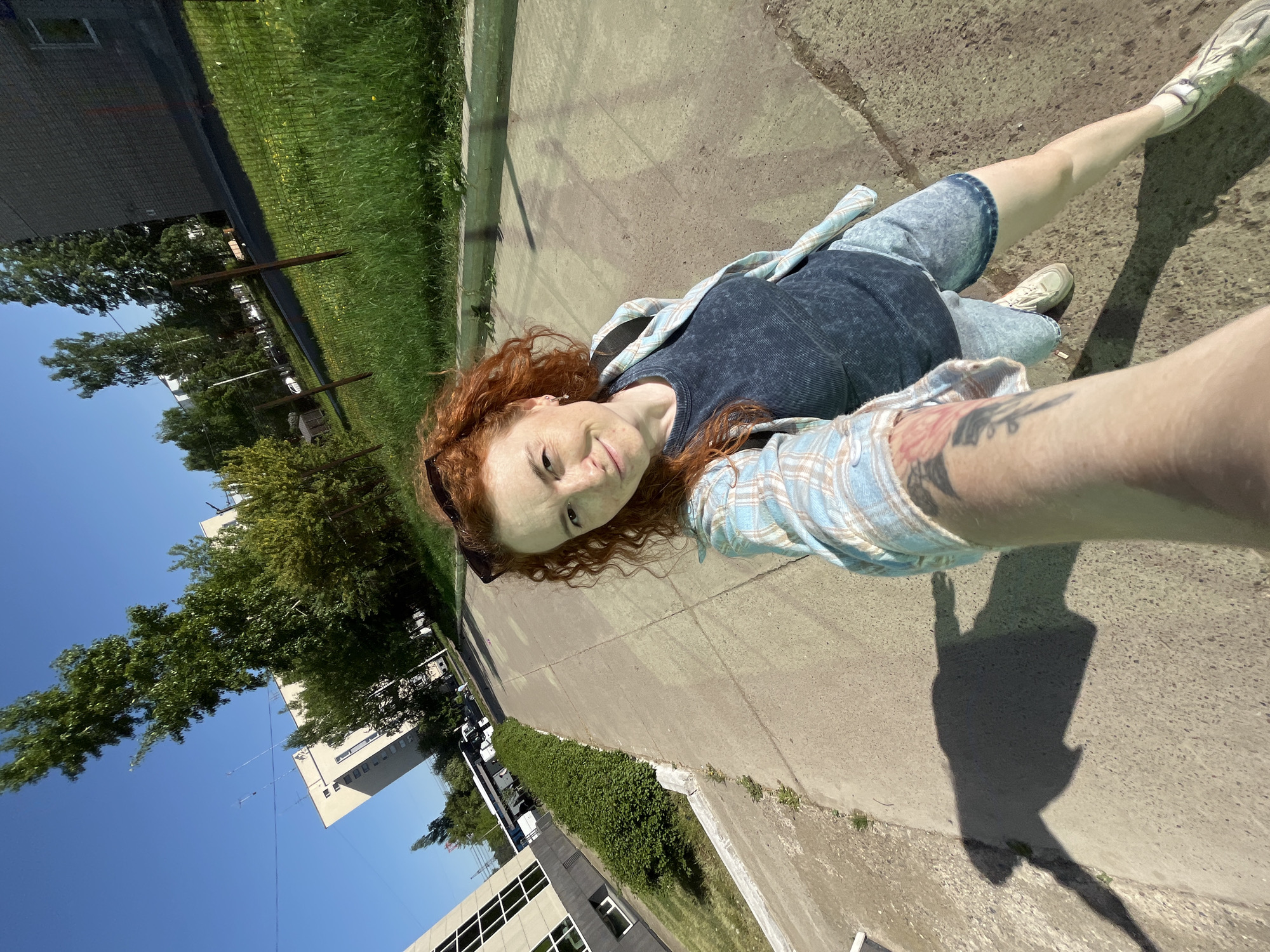 A young woman with red curly hair is taking a selfie in an urban setting. She is wearing a dark blue shirt, a light blue plaid shirt, and light blue shorts. Her arm is extended towards the camera, revealing a tattoo. The background includes a sidewalk, grass, trees, buildings, and a clear blue sky. The location appears to be near buildings and trees in Chaikovsky, Russia.