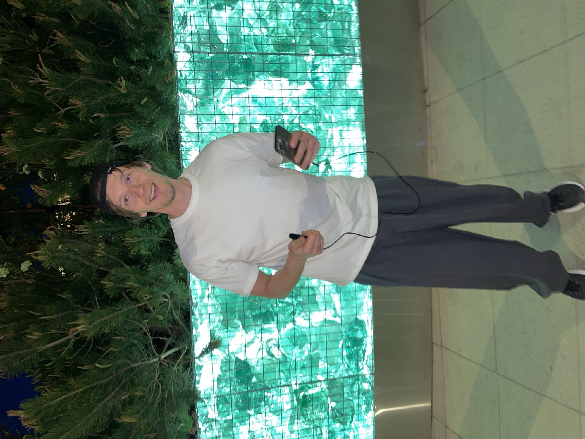 A man wearing a white t-shirt and dark pants stands in front of a green illuminated wall in what appears to be a public space. He is holding a smartphone in one hand and a small electronic device, possibly a microphone or remote, in the other, with a wire connecting to it. To the left of the man, there is a large green coniferous tree. The background suggests an interior setting with a tiled floor and diffused lighting. While Moscow, Russia is mentioned in the request, there are no indicators or signifiers suggesting this location.