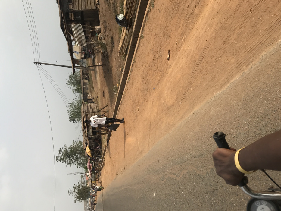 The scene depicts a street view in Ogbese, Nigeria, under daylight conditions. The foreground features the right hand and forearm of an individual operating a motorcycle or similar vehicle, gripping a black handlebar. A yellow wristband is present on the subject's left wrist. The immediate ground consists of an asphalt road surface transitioning to a reddish-brown dirt shoulder on the right.
In the midground, a dark-skinned male subject walks from left to right on the dirt shoulder. He wears a white t-shirt with a multi-colored graphic on the chest (four playing card suits), dark shorts, and dark footwear. He carries a black bag in his left hand. A distinct shadow extends from his left side onto the dirt. The dirt shoulder displays tire tracks and scattered debris. A concrete curb separates the paved road from the dirt shoulder in the midground.
The background reveals a series of rudimentary roadside structures, constructed primarily from wood and corrugated metal, consistent with market stalls or small shops. Some structures feature fabric awnings. A tall wooden utility pole with multiple overhead wires is visible on the right side. Piles of rough-cut wooden planks are stacked near one of the structures on the far right. Further down the road on the left, additional vehicles and indistinct figures are present. The sky above is light and hazy. No direct interactions between individuals are discernible.