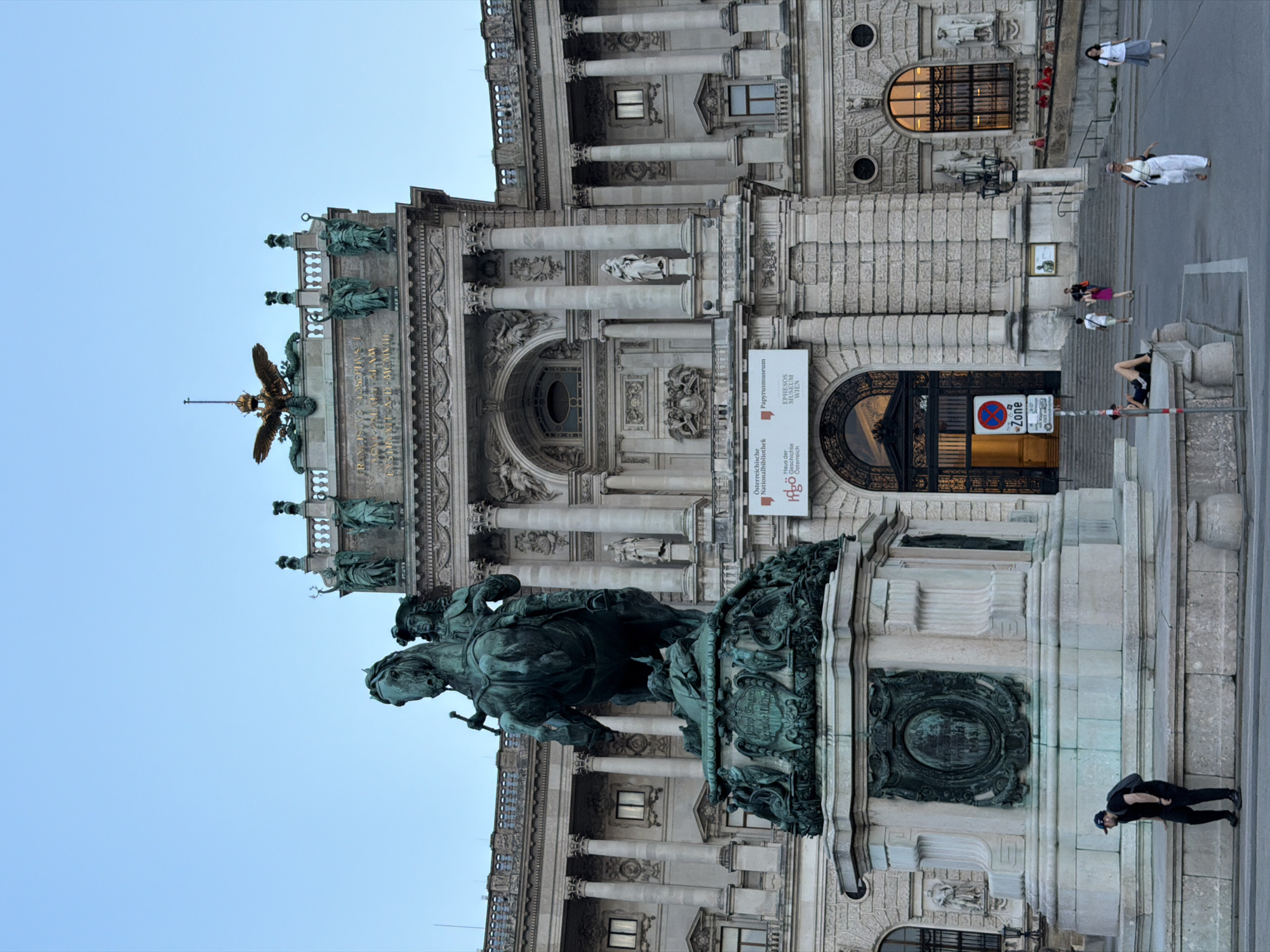 The image depicts the equestrian statue of Archduke Charles in front of the Neue Burg wing of the Hofburg Palace in Vienna, Austria. The statue, cast in bronze, is mounted on a large stone pedestal adorned with relief sculptures. A man wearing a black cap, black t-shirt, and black pants with a backpack is bending down near the base of the monument. In the background, the grand facade of the Neue Burg, a historical imperial palace, is visible with its classical architecture, columns, and statues. Several people are seen walking on the pavement and stairs in front of the palace, and some are seated on the steps. Banners advertising museums are displayed on the building's entrance. The sky is clear and light blue, suggesting it is either early morning or late evening.