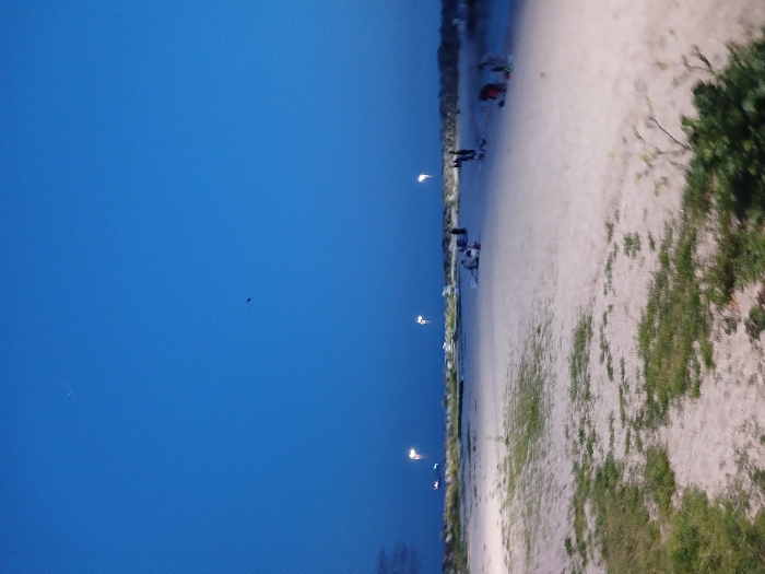 The image depicts a beach in Istanbul, Türkiye, at dusk. The coordinates (40.956, 28.816) correspond to a location along the coast of the Sea of Marmara, near the Florya neighborhood. The sky is dark blue, with a few stars visible. The beach is empty except for a group of people sitting on the sand in the distance. The sand is white and there is sparse vegetation growing on it. Several streetlights are visible in the background, illuminating the scene. The image was likely taken during the evening hours, as the sky is still light but the sun has set.