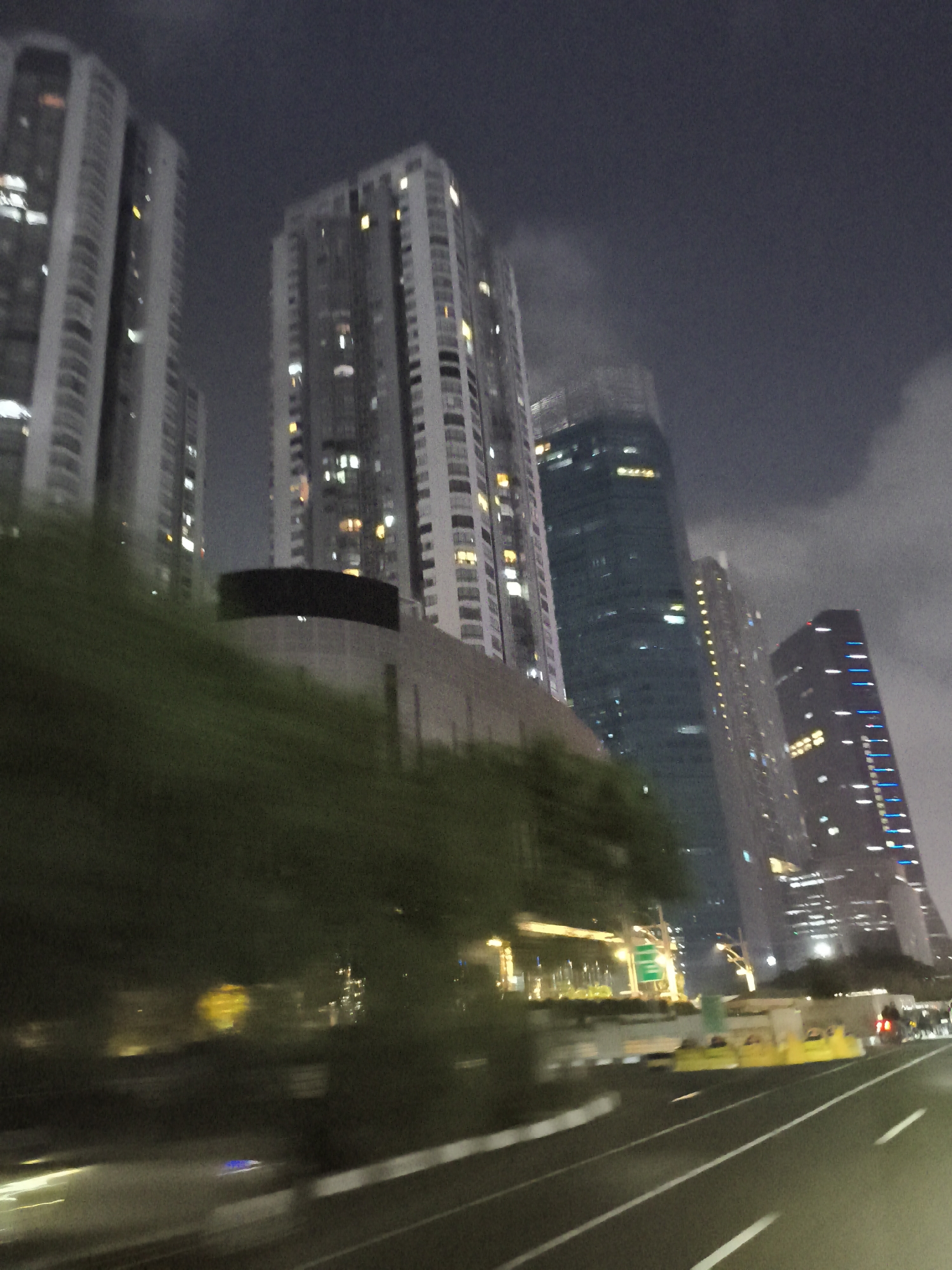 The image depicts a nighttime view of a highway in West Jakarta, Indonesia, with a line of tall, illuminated skyscrapers in the background. The buildings vary in design, with some featuring uniform rows of windows and others with more distinct architectural elements. The sky is dark and cloudy, obscuring any stars or moon. The foreground shows a road with white lane markings, and the motion blur suggests the photograph was taken from a moving vehicle. Some trees and foliage are visible along the side of the road, and there are some yellow traffic barriers and street lights. The overall impression is one of a bustling urban environment at night.