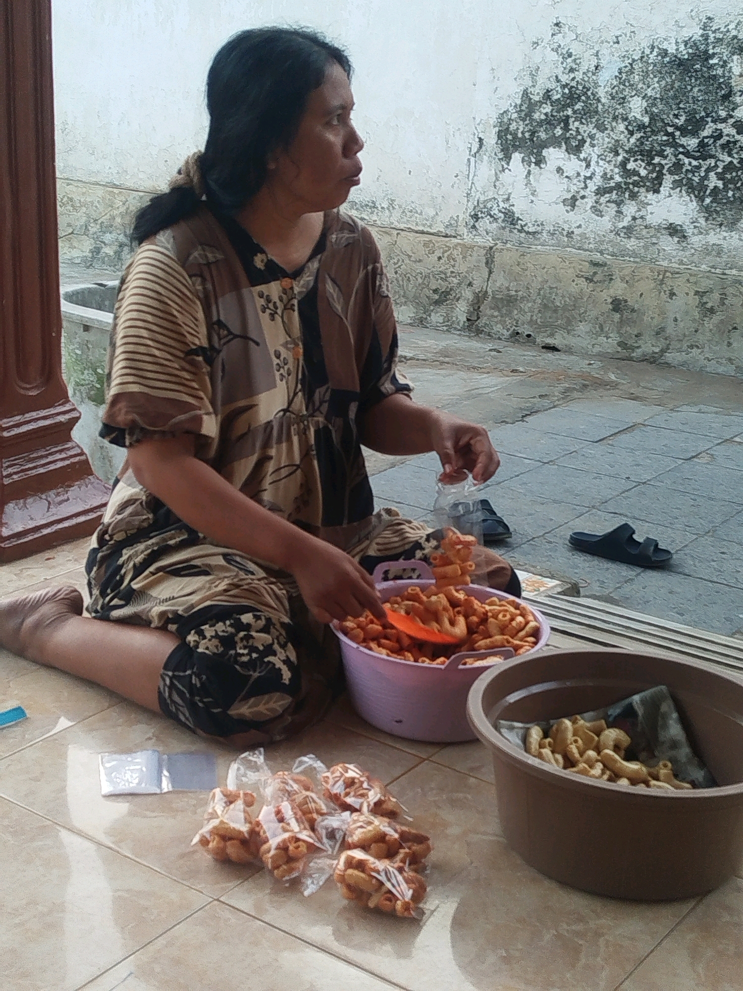 In Burneh, Indonesia, a woman is sitting on the tiled ground, facing right. She is wearing a patterned dress with a floral and striped design. Her right hand is reaching into a large purple bowl filled with snacks. In front of her are several small, clear plastic bags containing more snacks. To her right, a dark brown bowl holds a pile of snacks, some of which are wrapped in a dark fabric. Her left leg is bent, and her right leg is extended in front of her. The background consists of a textured, off-white wall with dark stains, and a tiled floor. Two dark sandals are visible on the floor behind her. A brown pillar is to the left of the frame.