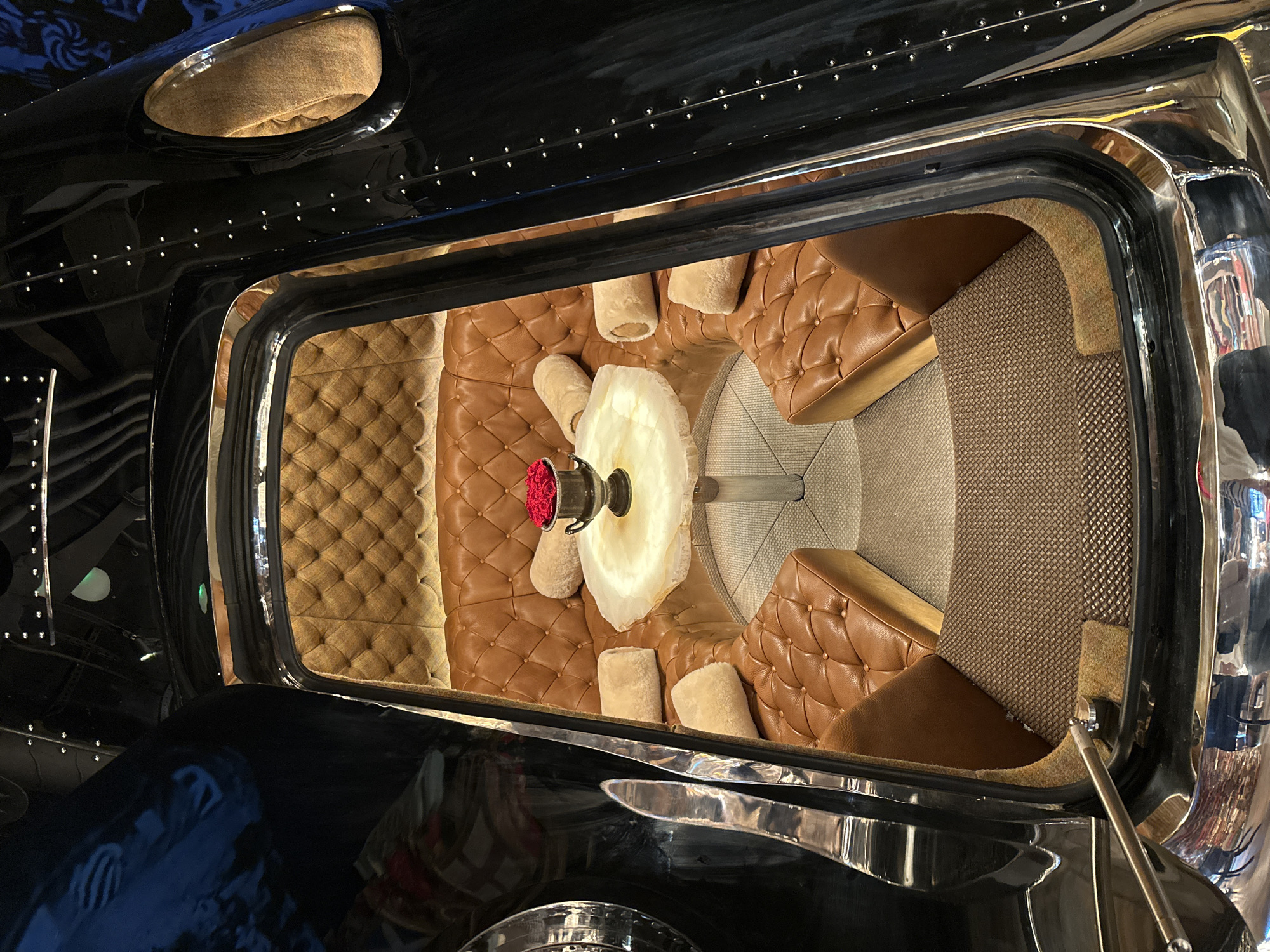 The image displays the interior of a luxurious, custom-designed seating area, possibly within a vehicle or a themed establishment. The space is characterized by deep, button-tufted brown leather upholstery, forming a curved couch and individual seats. A small, translucent table with a rough, natural edge sits in the center, adorned with a silver vase holding a bouquet of red roses. Round, fluffy pillows are scattered across the seating. The exterior of this enclosed space is glossy black, with visible metallic rivets and curved metallic trim, suggesting a high-end, bespoke construction. The flooring is a textured, dark grey carpet. The overall aesthetic evokes a sense of opulent privacy and retro-futurism. The location is identified as London, United Kingdom. There are no people visible in the image.