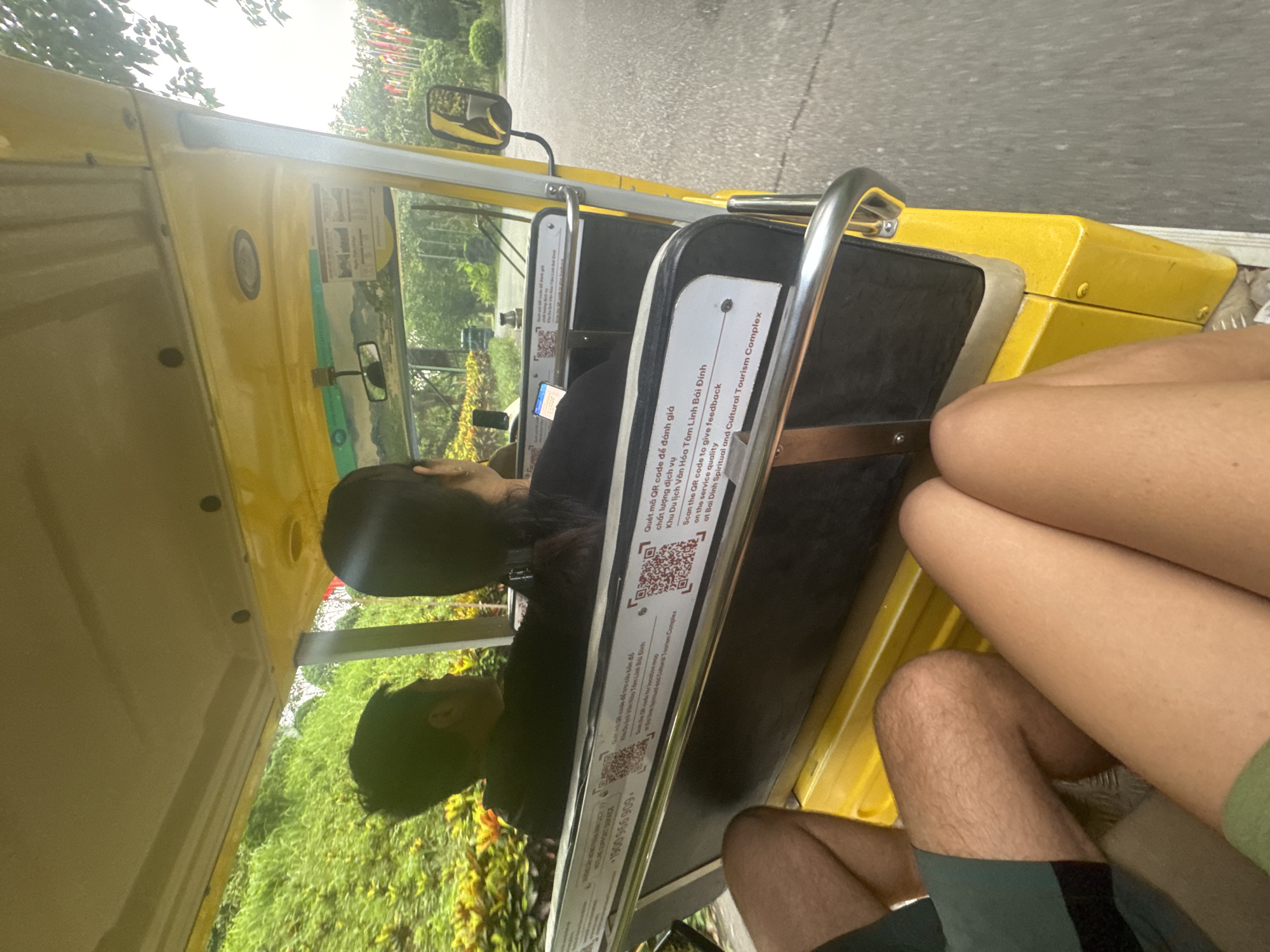 In the city of Tây Hoa Lư Ward, Vietnam, a yellow open-air electric vehicle, likely a tourist buggy, is captured in motion on a paved road. The vehicle's interior is visible, revealing a driver and a passenger seated in front, both facing forward. The driver, a person with dark hair, is operating the vehicle, while the passenger, also with dark hair, is looking ahead. In the foreground, the legs of two individuals are visible, suggesting they are passengers in the rear of the buggy. One person wears dark green shorts, and the other wears a green garment. The buggy has a white panel affixed to its rear, displaying QR codes and text in both Vietnamese and English, indicating a feedback mechanism for the "Bai Dinh Spiritual and Cultural Tourism Complex." The surrounding environment features lush green foliage, including trees and bushes, indicating a natural or park-like setting. The sky appears overcast. The overall scene conveys movement and a tourist experience within a scenic, likely cultural or spiritual, destination.