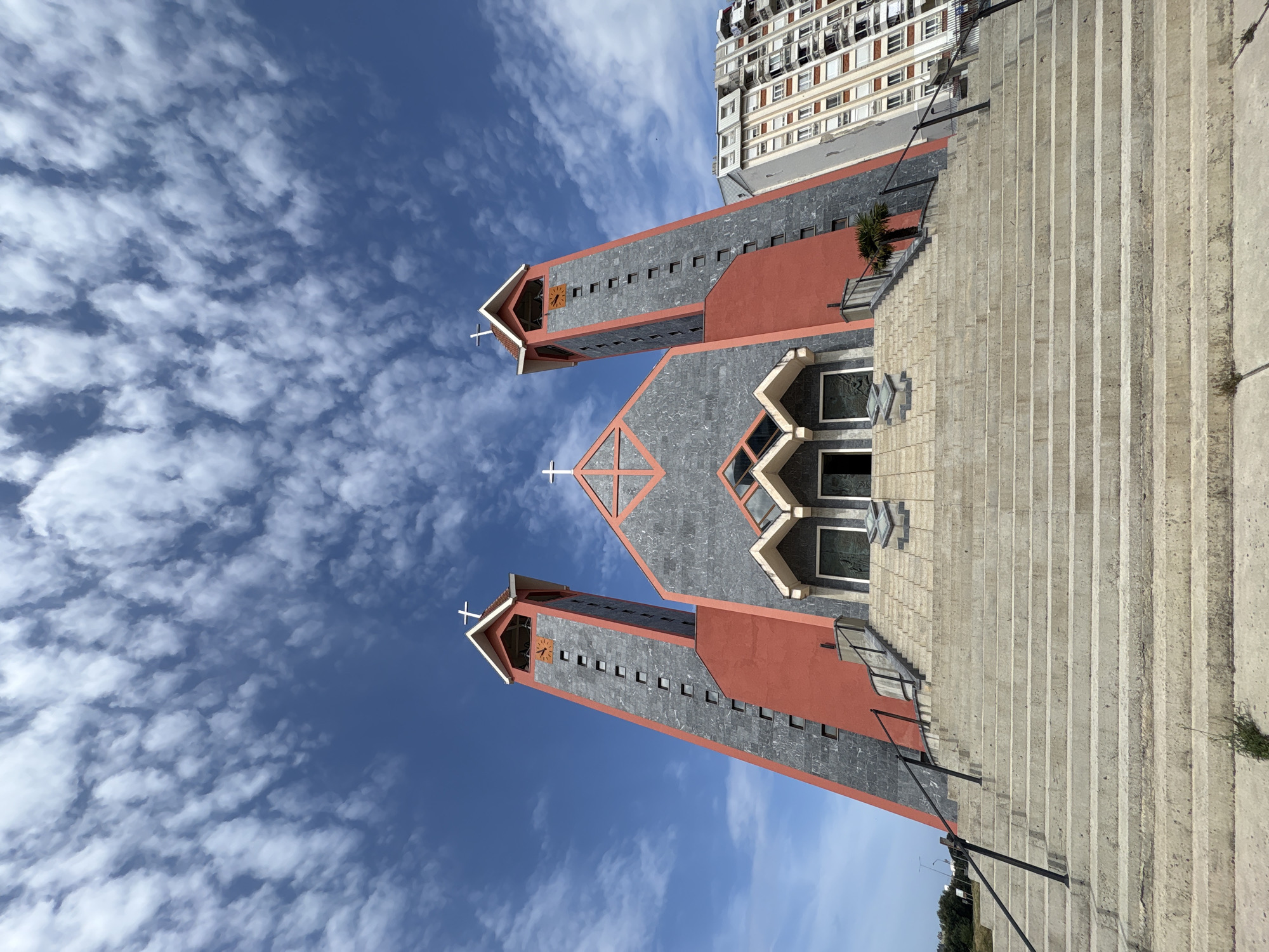 The image captures the imposing Roman Catholic Cathedral of St. Peter the Apostle in Bar, Montenegro, set against a vibrant blue sky filled with scattered clouds. The cathedral is constructed with a modern architectural design, featuring two prominent towers and a central structure. The exterior is a combination of gray stone and red plastered surfaces. A grand staircase leads up to the entrance of the cathedral. To the right of the church, a tall apartment building is visible. In the distance, green vegetation can be seen, giving context to the cathedral's location within the city.