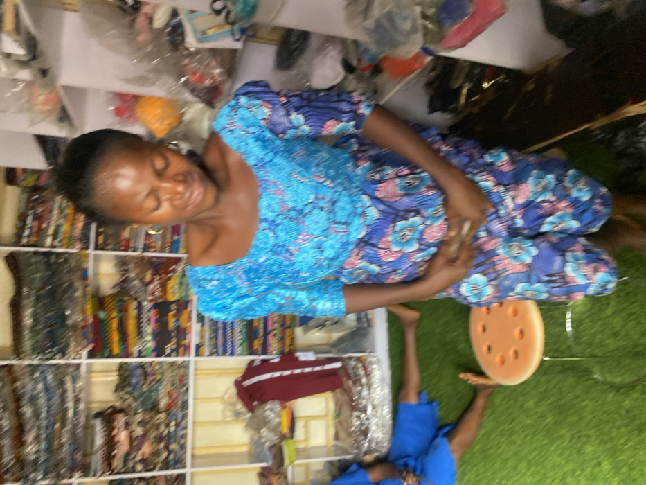 A woman sits cross-legged in a patterned blue dress. Behind her are shelves filled with rolls of colorful fabrics. The woman's hands are clasped in her lap, and she appears to be smiling. Below, another person is seated on the floor with their legs visible, wearing a blue outfit. Artificial grass is visible in the background.