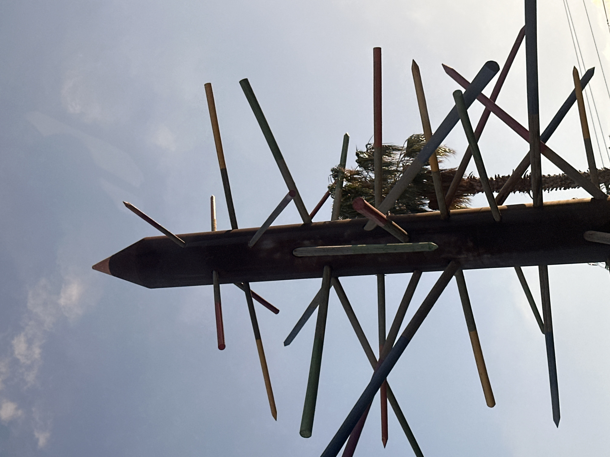 The image shows an outdoor art installation resembling a giant pencil with numerous colored dowels or sticks protruding from its sides at various angles. The installation is positioned against a bright, slightly cloudy sky. The central object is a tall, dark, cylindrical structure with a conical top, mimicking the shape of a pencil. The colored sticks, which appear to be made of wood and painted in hues of red, yellow, green, blue, and white, are inserted into holes or slots along the main structure. Some of the sticks have sharpened points, resembling pencil tips. In the background, the fronds of a palm tree are visible, suggesting a warm climate. The overall impression is one of a whimsical, large-scale sculpture. This art installation is located in the city of Petah Tikva, Israel.