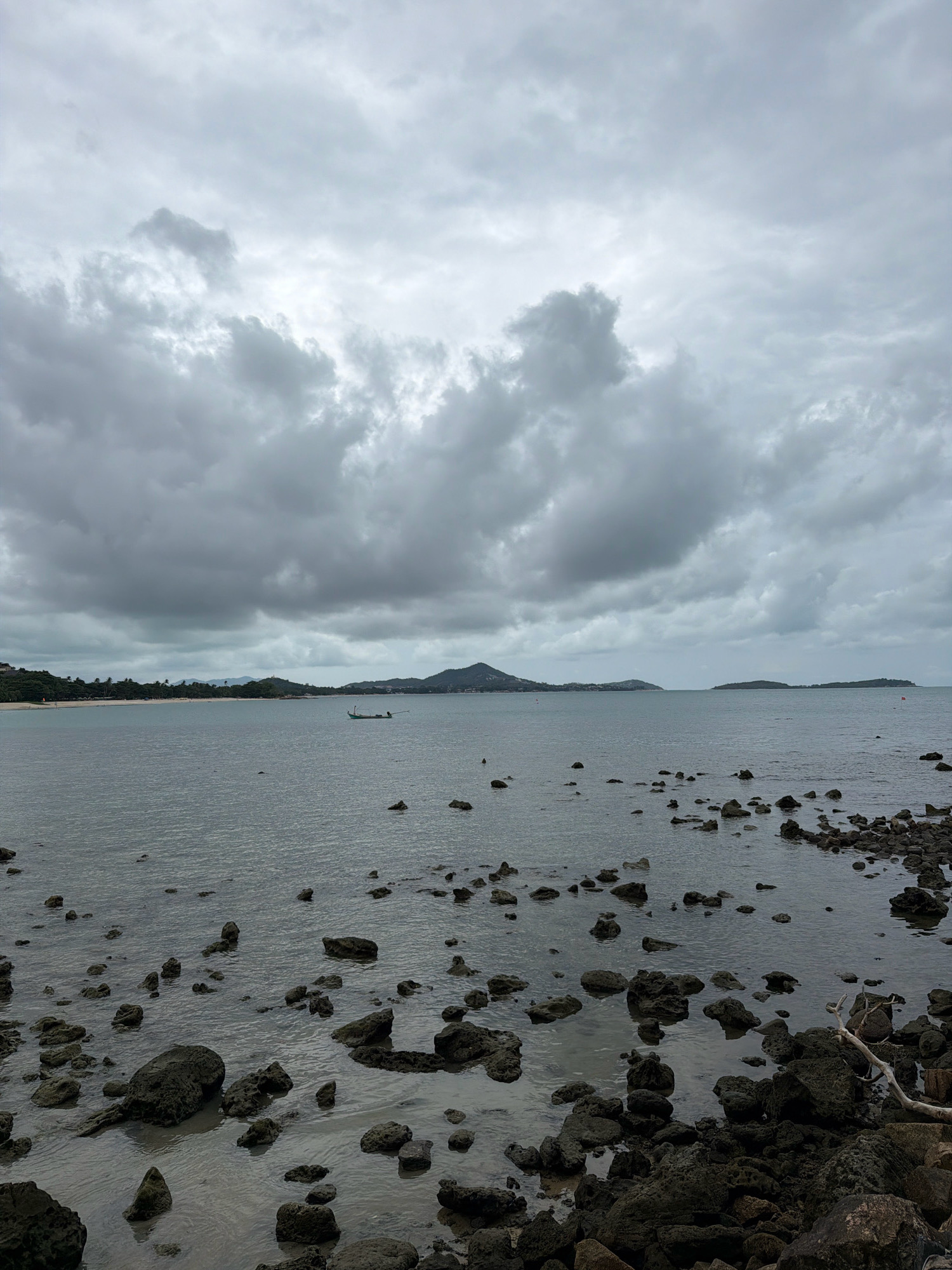A full shot captures the rocky shoreline and the calm sea under a cloudy sky in the Ko Samui District of Thailand. In the foreground, dark, wet rocks dot the water's edge, with some submerged and others exposed. The sea is a muted gray, reflecting the overcast sky. In the mid-distance, a small, green fishing boat with a long wooden pole floats on the water. Beyond the boat, a long stretch of coastline is visible, featuring a sandy beach with scattered palm trees and lush green vegetation. Hills and mountains form the backdrop, disappearing into the hazy sky. The clouds are heavy and gray, suggesting impending rain or a continuation of overcast weather. No people are clearly visible in the frame.