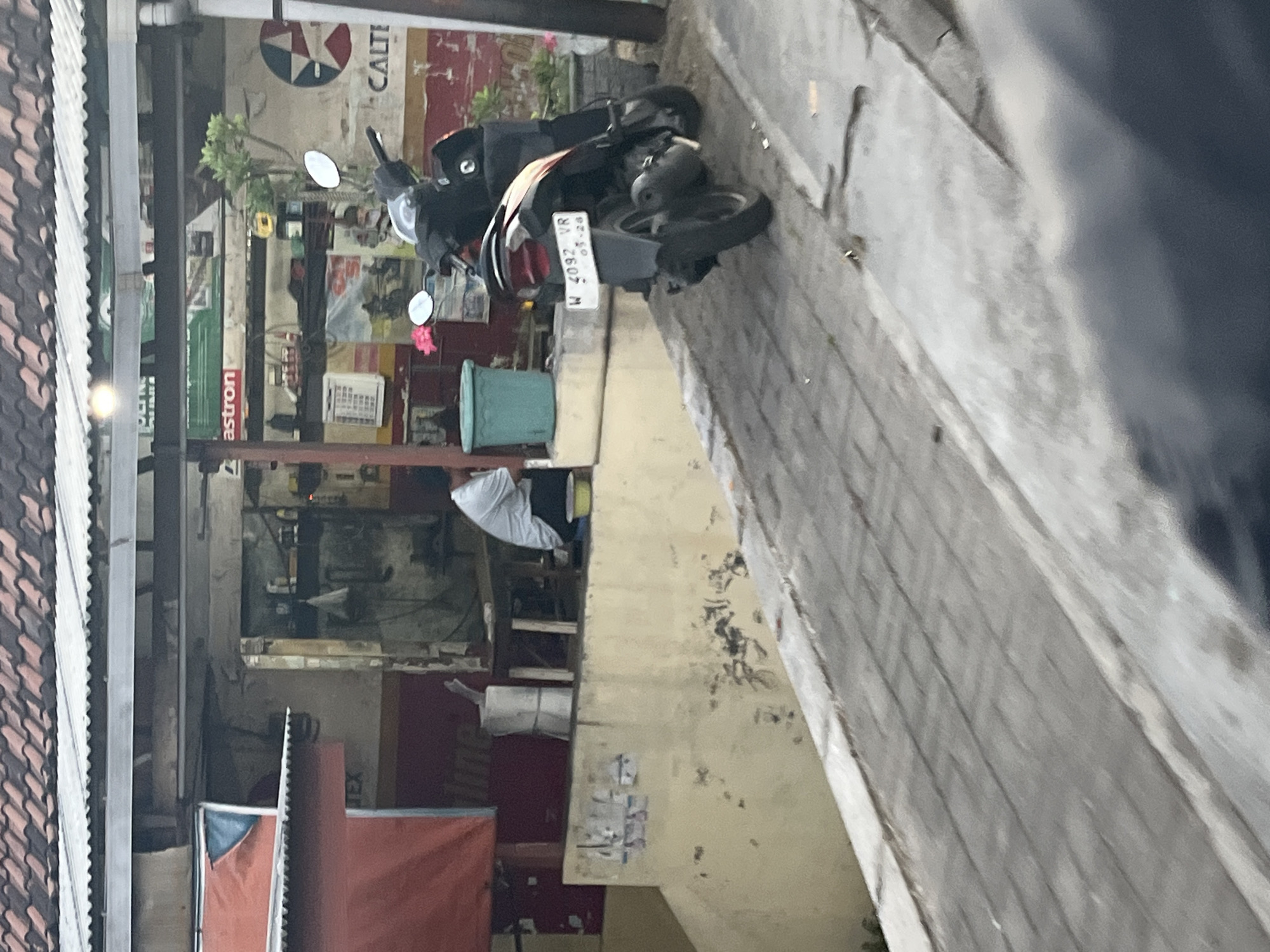 The image depicts a roadside scene in Kludan, Indonesia, in front of a workshop. A dark-colored scooter with a license plate reading "W 4092 VR 03-28" is parked on a sloped, paved walkway. To the left of the scooter, a man in a white shirt is seated on the ground next to a turquoise bucket. Behind the man, a workbench is visible, cluttered with tools and various items. Above the workbench, shelves display more items, including a white box, bottles, and what appears to be a framed picture. A large red tarp covers a section of the building to the left. The building itself has a corrugated metal roof. The background includes a wall with a faded logo featuring a star within a circle, and the word "CALT" is partially visible. The environment appears to be an outdoor urban setting with a worn, textured surface for the walkway.