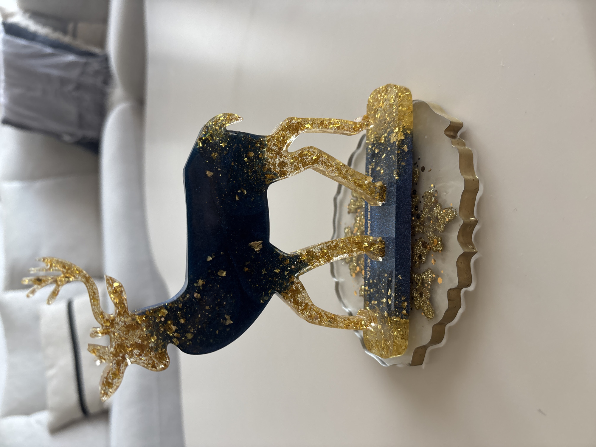 A decorative resin deer figurine, predominantly dark blue with gold flakes, stands on a clear and gold-flecked base with a wavy edge. The figurine is positioned in the foreground, casting a slight shadow on a white, flat surface. In the blurred background, a white couch with cushions and a grey throw is visible. The scene is indoors, with ample natural light. There are no people or animals present. The location is not specified to be Dubai, United Arab Emirates.