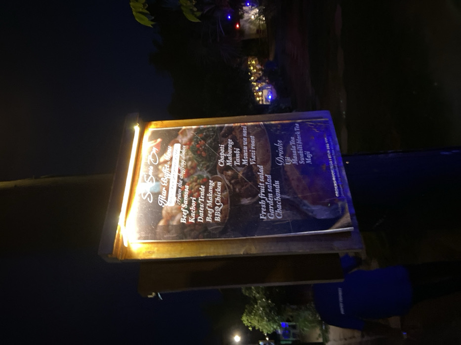 The image shows a menu board illuminated by a strip of light above it. It is mounted on a wooden post and is positioned on a sidewalk at night. The menu is for a restaurant called "Spice Bay." It advertises a "flair buffet menu" of various dishes, including beef samosa, kachori, dates/tende, beef makange, BBQ chicken, chapati, maharage, tambi, meusu wa nazi, viazi roast, fresh fruit salad, garden salad, chachandu, uji, masala tea, Swahili black tea, and maji. The image also shows a person in a blue shirt walking away from the menu, passing on the left side. This scene is located in Dar es Salaam, Tanzania, at the coordinates -6.754, 39.248.