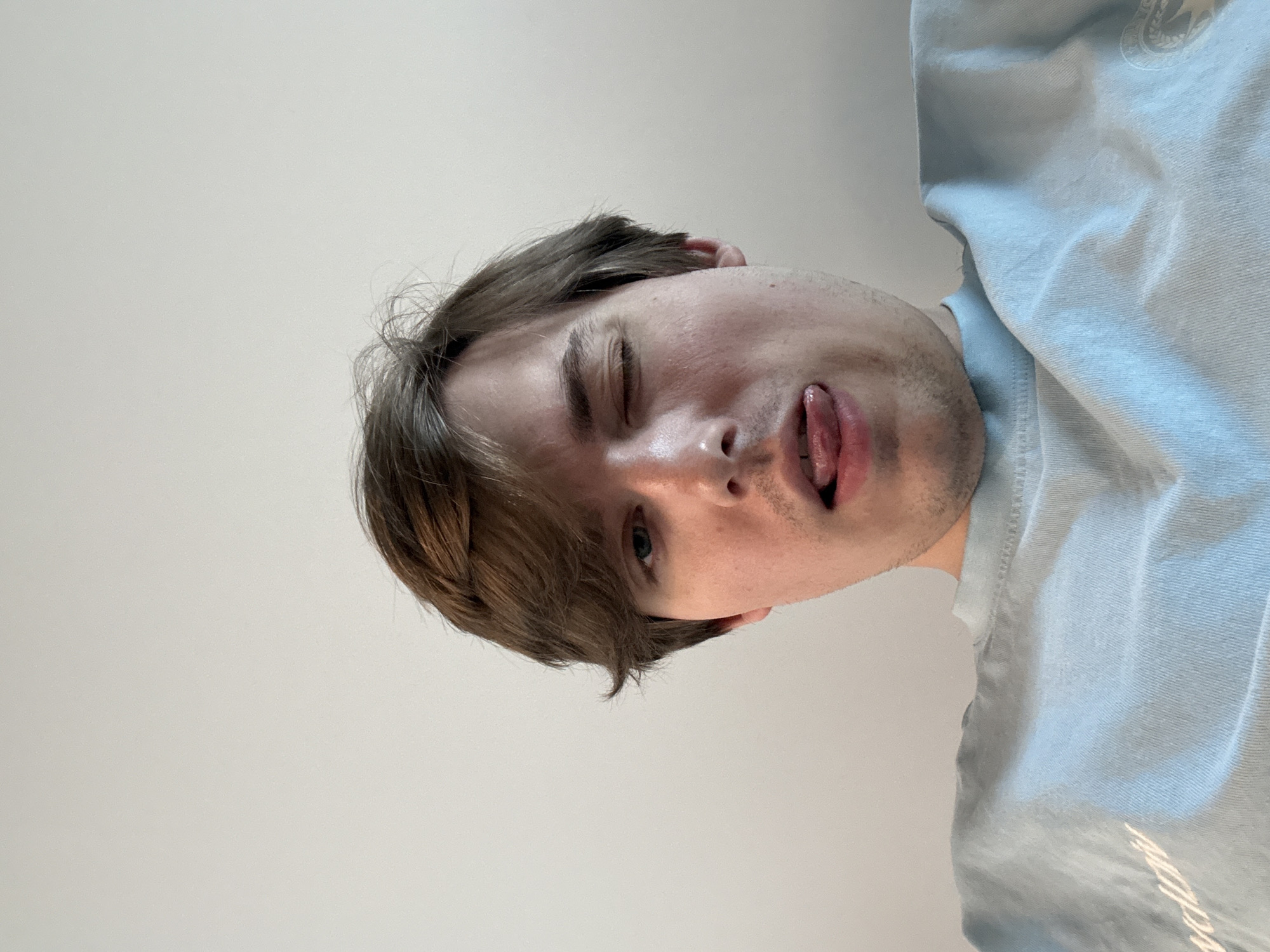 The image shows a close-up, low-angle, selfie-style shot of a young man with light brown hair and blue eyes. He is winking one eye and sticking his tongue out. He is wearing a light blue t-shirt. The background is a plain, light-colored wall. There is no discernible information about the location in Dnipro, Ukraine.