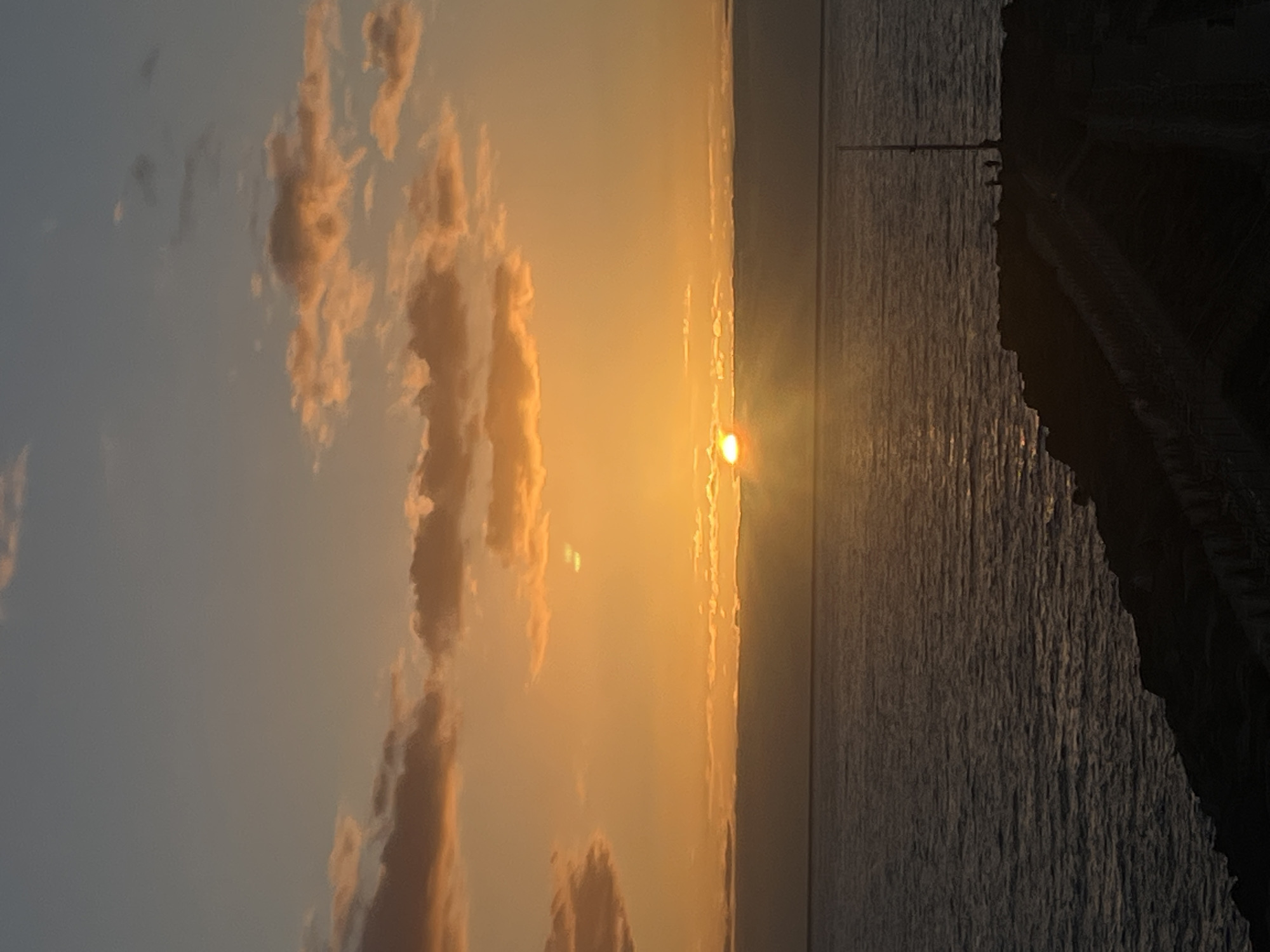 The image depicts a sunset over the ocean. The sky is a mix of orange, gray, and light blue with clouds. The sun is visible as a bright orange orb on the horizon. The ocean is reflecting the sunlight, creating a sparkling effect. A dark, rocky shoreline is visible on the right side of the image. The image was taken in Mogán, Spain.