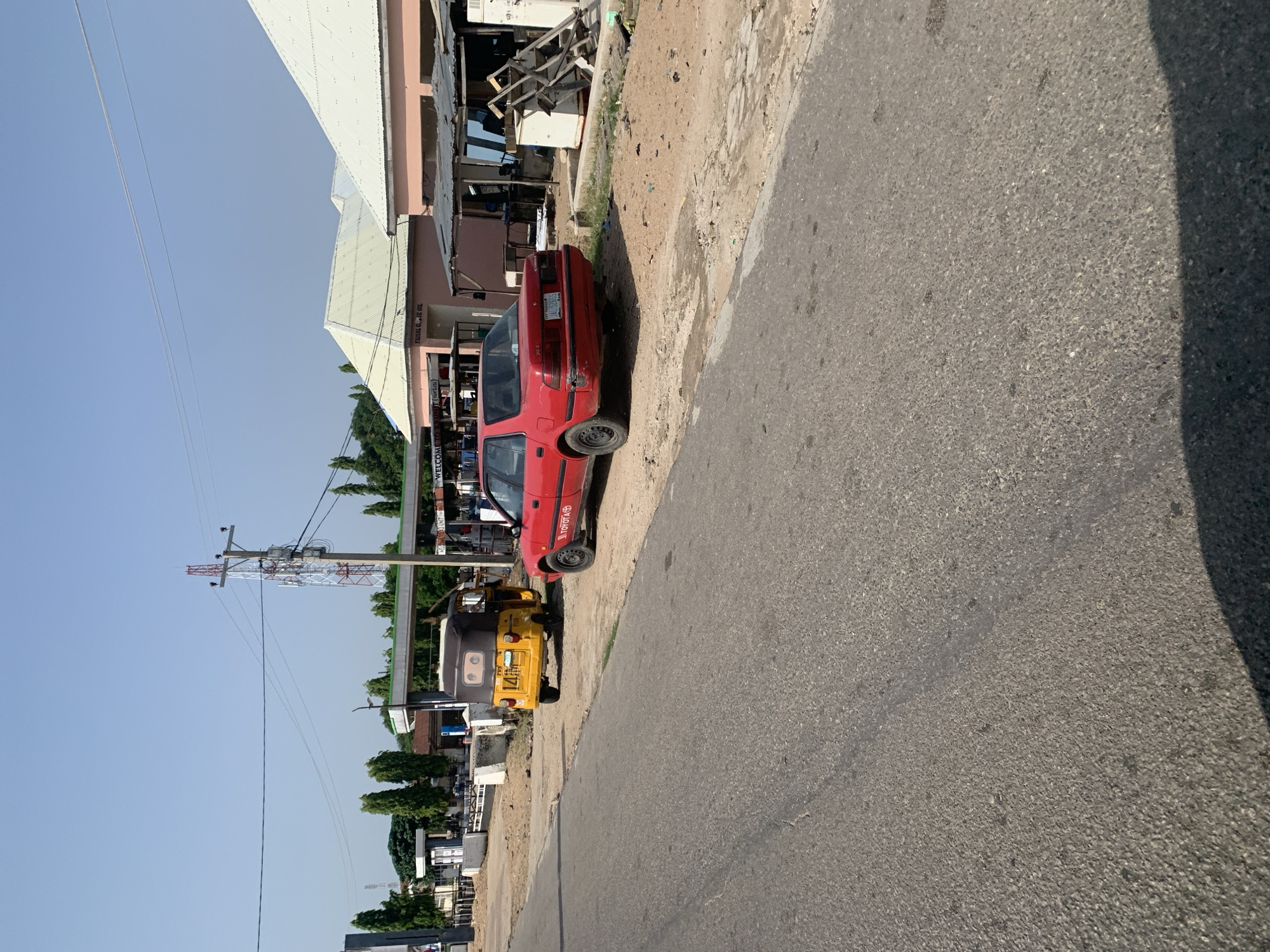 In an outdoor, daytime scene, a red Toyota sedan is parked on the side of a paved road. To the left of the sedan, a yellow auto-rickshaw is also parked. Behind the vehicles, there are several buildings with corrugated metal roofs and pastel-colored facades. A gas station canopy is visible between some of the buildings. The area appears to be a commercial strip with a few trees scattered around. The sky is clear and blue. The overall environment suggests a sunny day in a town or city.