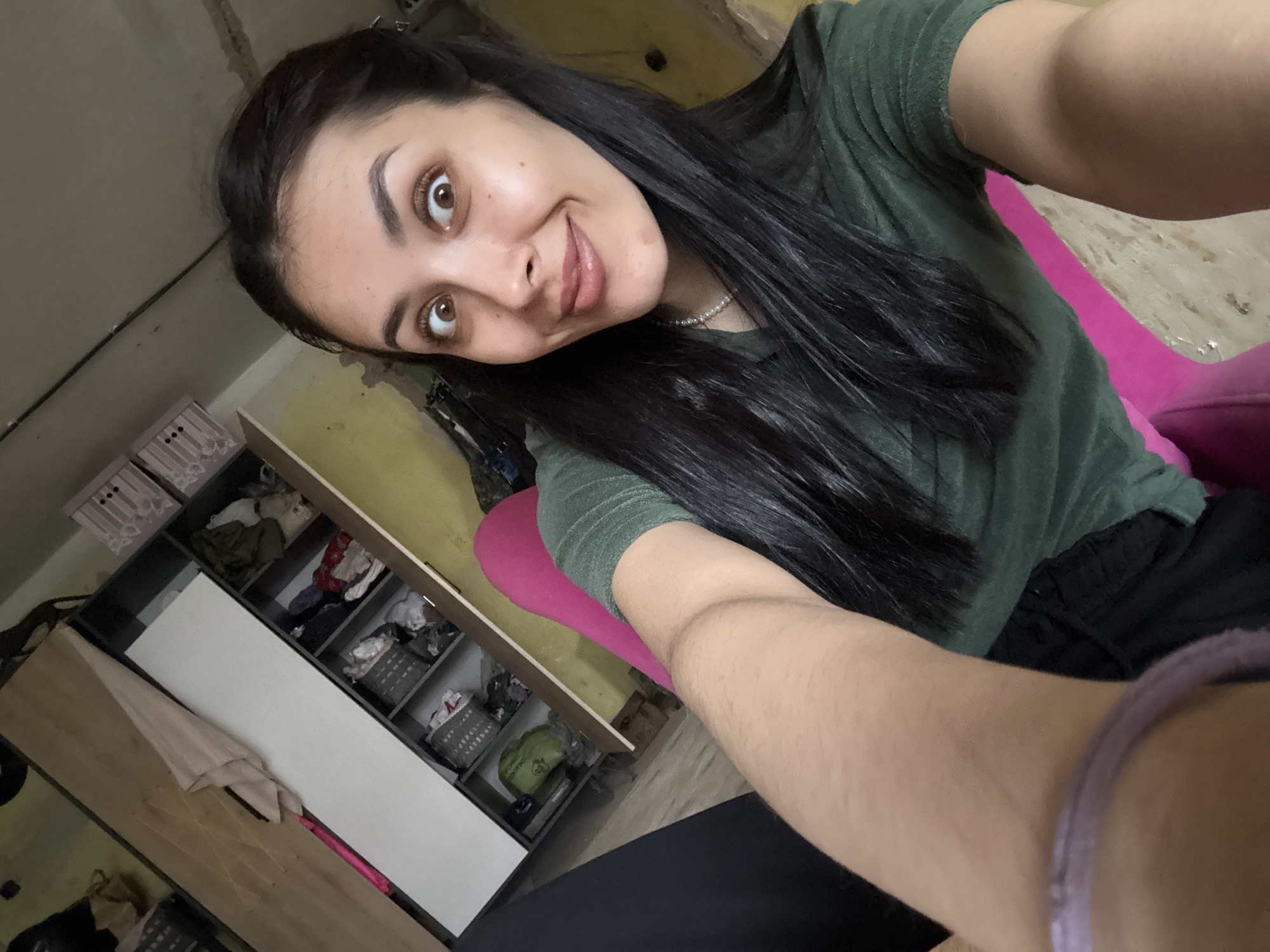 A young woman is taking a selfie in a room. She has long, dark hair, brown eyes, and is wearing a green t-shirt and a pearl necklace. She is sitting in a pink chair. Behind her is an open closet with clothes and storage boxes. The walls are painted yellow. The location is not identifiable as Votkinsk, Russia from the image.