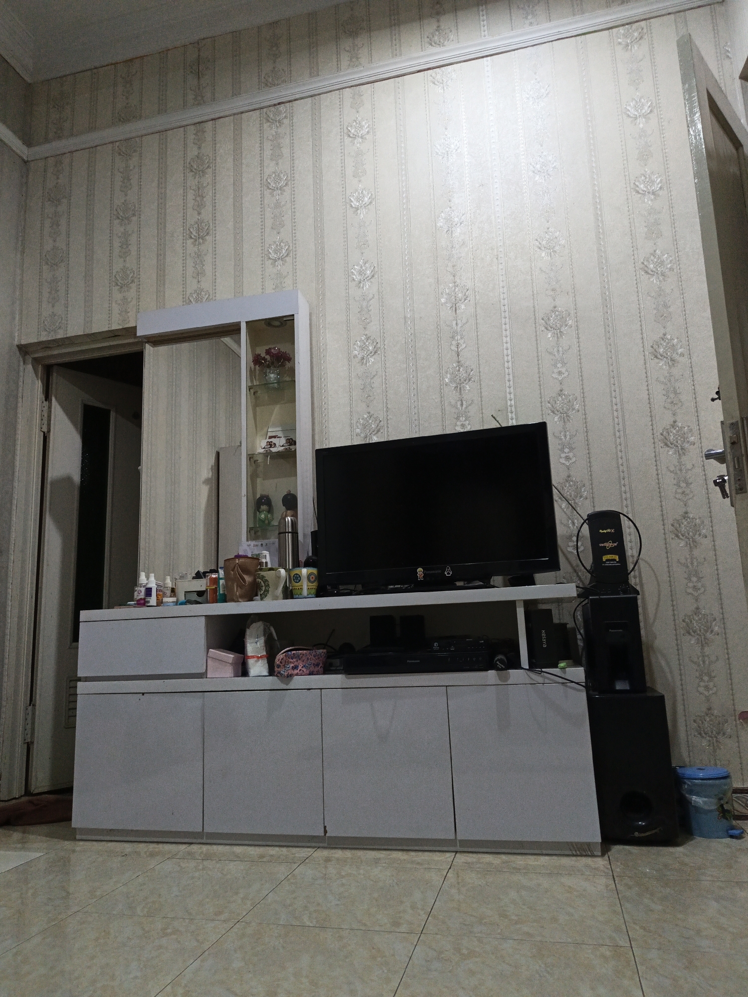 The image shows an indoor scene, likely a living room, located in Karangpakel, Indonesia. The room features a light-colored tiled floor and walls covered with patterned wallpaper. In the center of the frame, there is a white entertainment console with a black television on top. To the left of the television, there is a mirrored cabinet with shelves displaying various items, including a vase of red flowers, figurines, and a stainless steel thermos. On the console itself, there are several bottles and containers, suggesting personal care items. To the right of the television, a black speaker sits on the floor. The room appears to be well-lit, with the wallpaper reflecting the light. There are no people visible in the image.