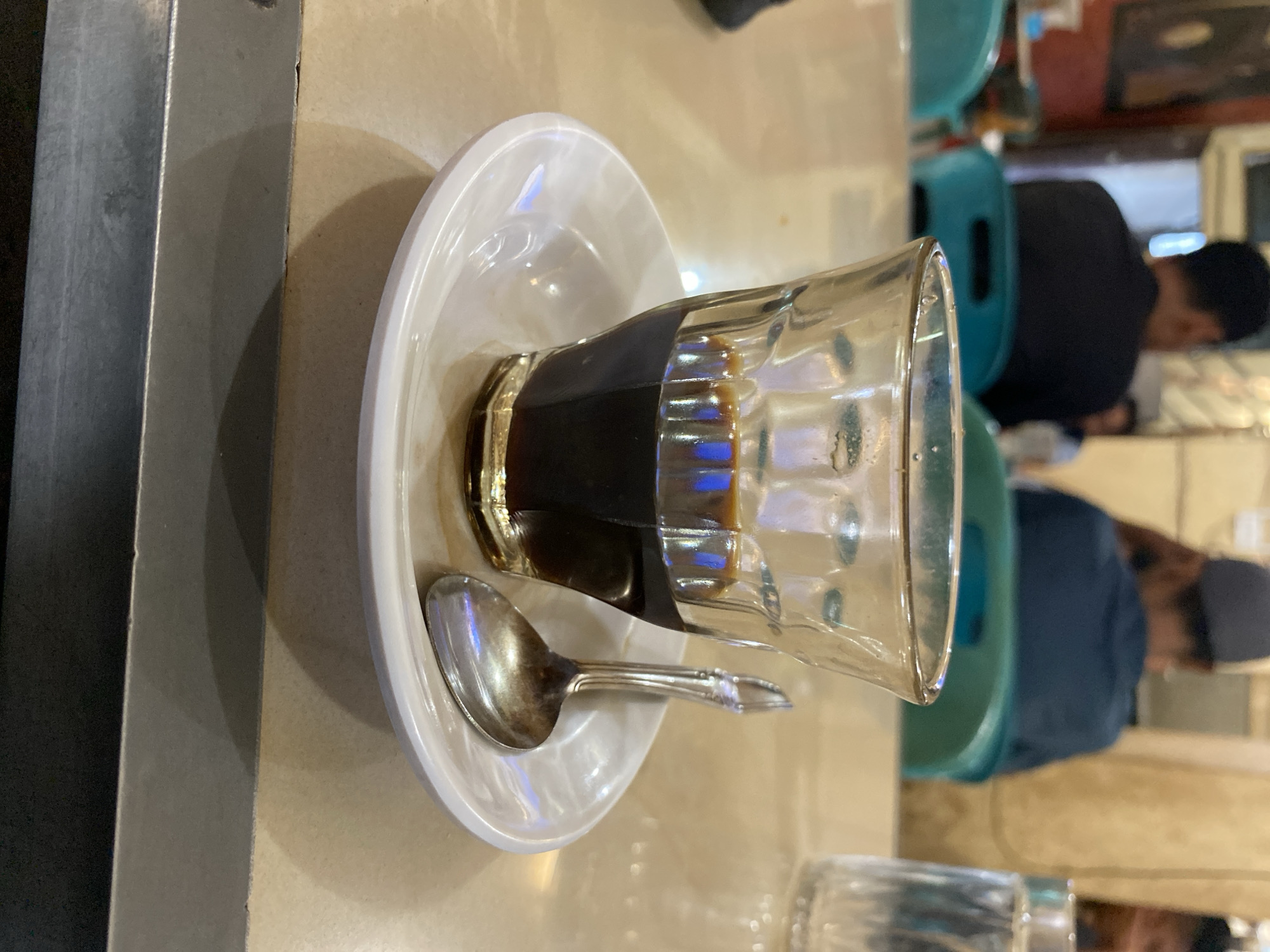 The image shows a close-up view of a glass of dark coffee on a white saucer, with a small silver spoon resting on the saucer. The coffee is dark brown and fills about a third of the glass. The glass has a textured surface and is transparent, allowing the reflection of blue and white light from above. The saucer is white and circular, with a slightly raised edge. The spoon is also silver and appears to be slightly tarnished.
The scene is set on a table with a light-colored, speckled surface, possibly a countertop or a table in a cafe. In the background, blurred figures of people are visible, seated at tables. The person closest in the background to the left is wearing a dark shirt. To the right, another person is wearing a dark blue shirt and a grey cap. There are also hints of green and turquoise chairs in the mid-ground, suggesting a public seating area. A glass of water is partially visible in the far right background.
The overall ambiance suggests a casual cafe or eatery, likely a local establishment. Considering the reference to Lhokseumawe, Indonesia, this scene could depict a coffee shop within the city, where locals gather to enjoy coffee. The setting appears to be indoors, with artificial lighting contributing to the reflections in the coffee glass. The image is focused on the coffee and its accompaniments, with the background providing context.