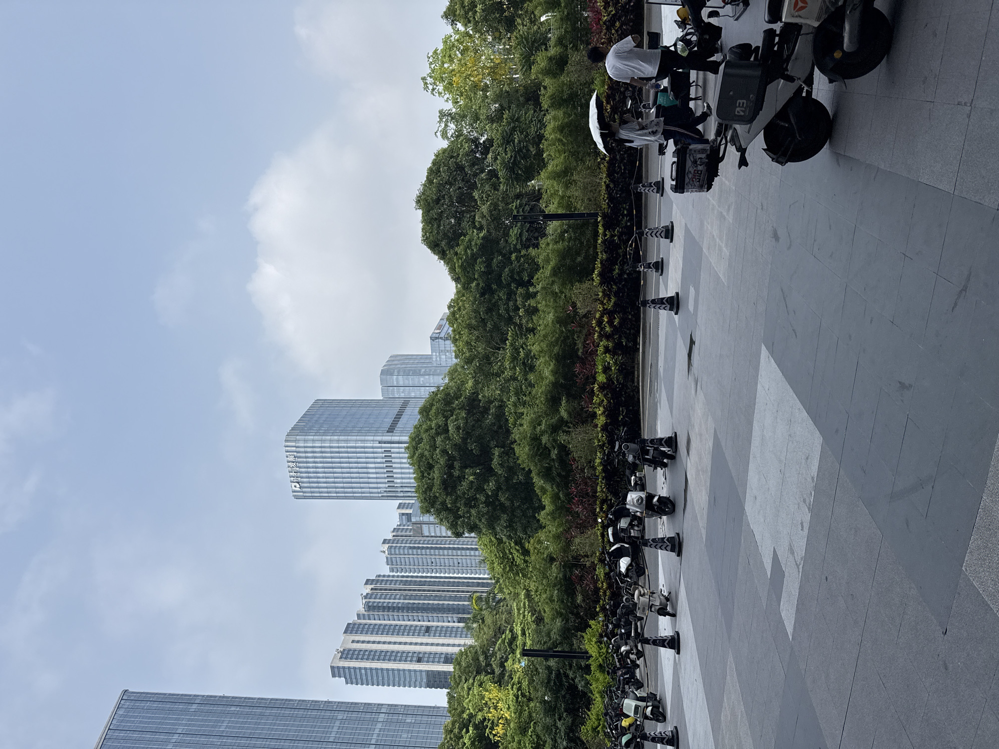 This image depicts an outdoor scene in Tianhe District, China, featuring a row of parked electric scooters and motorcycles in the foreground. In the middle ground, a dense line of lush green trees forms a natural backdrop, obscuring the lower portions of several tall, modern buildings that rise into the sky. Two individuals are walking in the mid-ground, one holding a white umbrella. The ground is paved with large, grey stone tiles. The sky is overcast with scattered clouds.