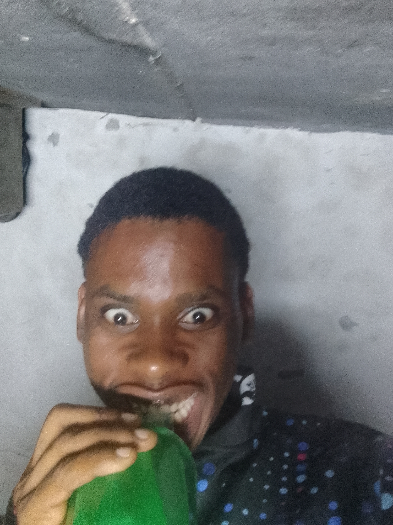 A person with dark skin and short black hair is holding a green bottle to their mouth with their right hand, contorting their face with wide eyes and an open mouth exposing teeth. The person is wearing a dark shirt with blue and white polka dots. The background is a grey, textured wall and ceiling. The location is the city of Unknown, Unknown.