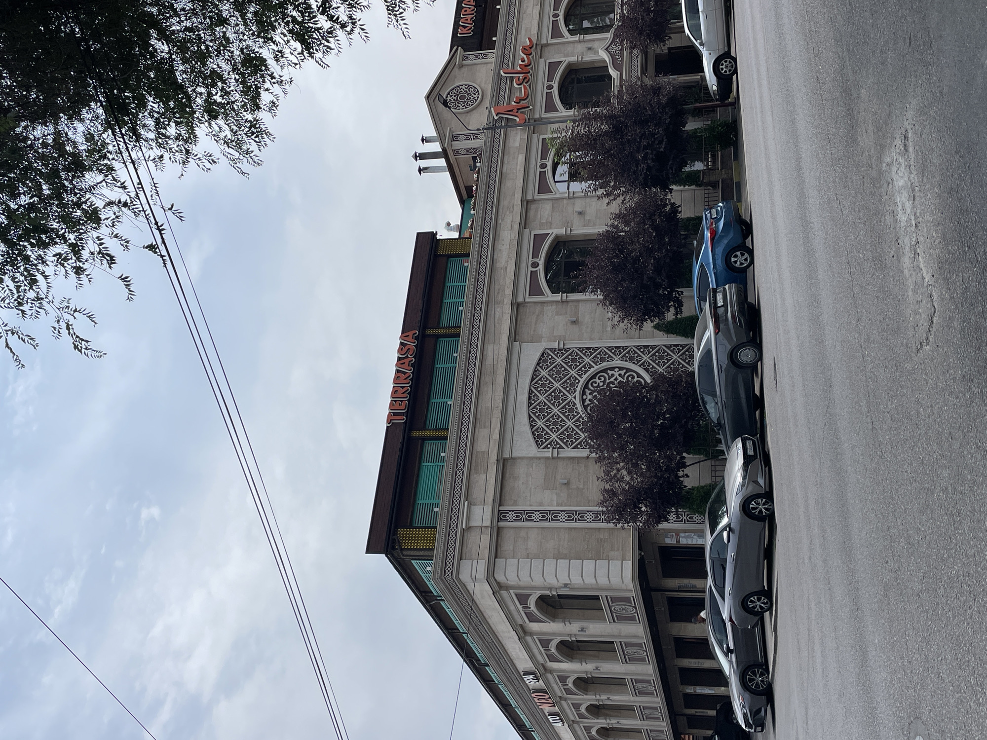 The image shows a street scene in Almaty, Kazakhstan, featuring a multi-story building with a restaurant or entertainment venue called "TERRASA" and "Arsha" visible on its facade. Several cars are parked along the street in front of the building. The architecture of the building appears ornate, with decorative elements and arched windows. Trees with dark foliage are planted along the sidewalk. The sky is overcast, and power lines are visible crisscrossing the upper portion of the frame. A person is seen standing near the entrance of the building. The overall scene depicts a typical urban environment on a cloudy day.