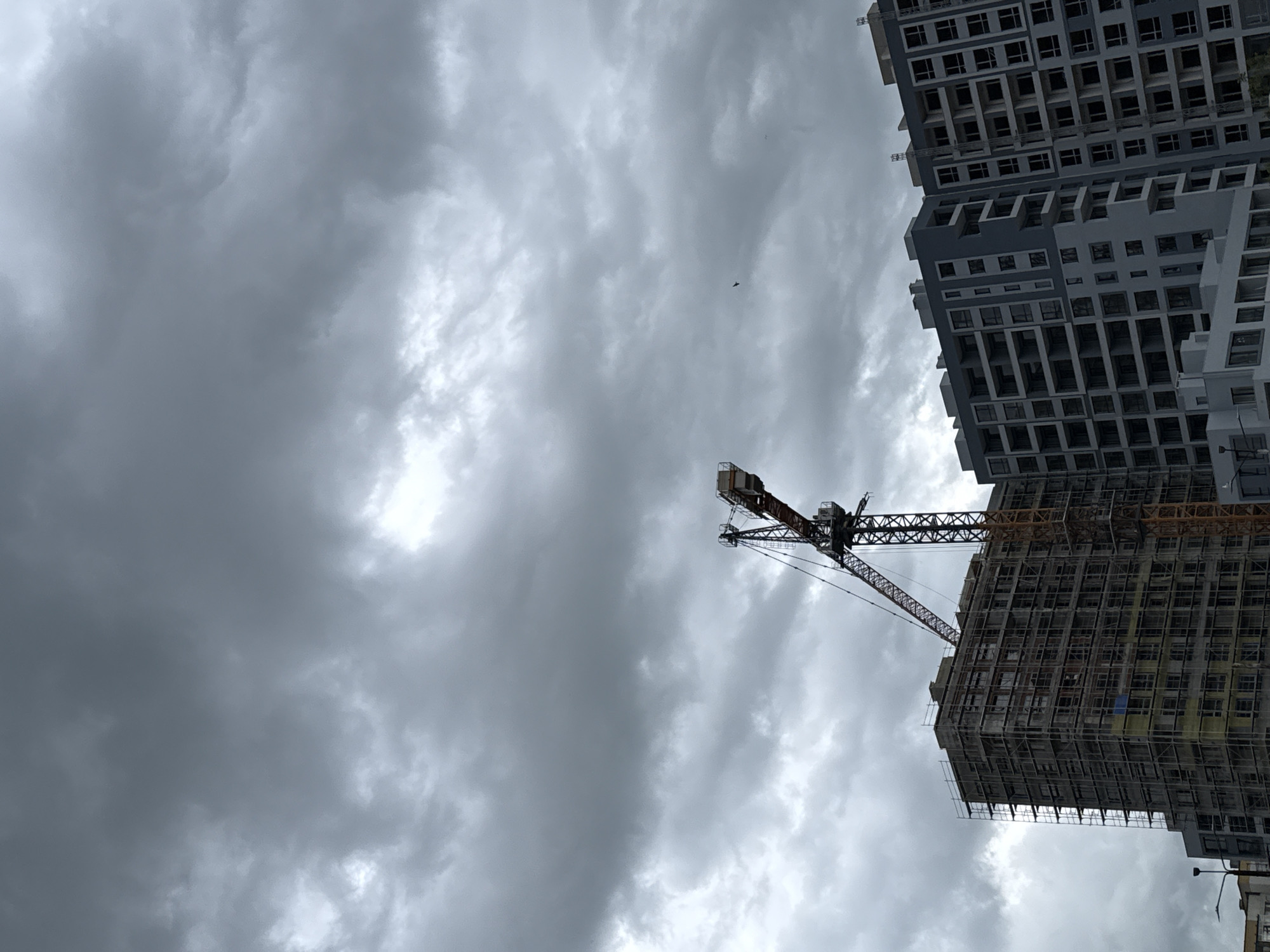 The image shows a construction site in an urban environment. A tall building under construction with scaffolding is visible on the right side, accompanied by a construction crane extending towards the left. Another completed high-rise building stands adjacent to the construction site. The sky is overcast with grey, heavy clouds.