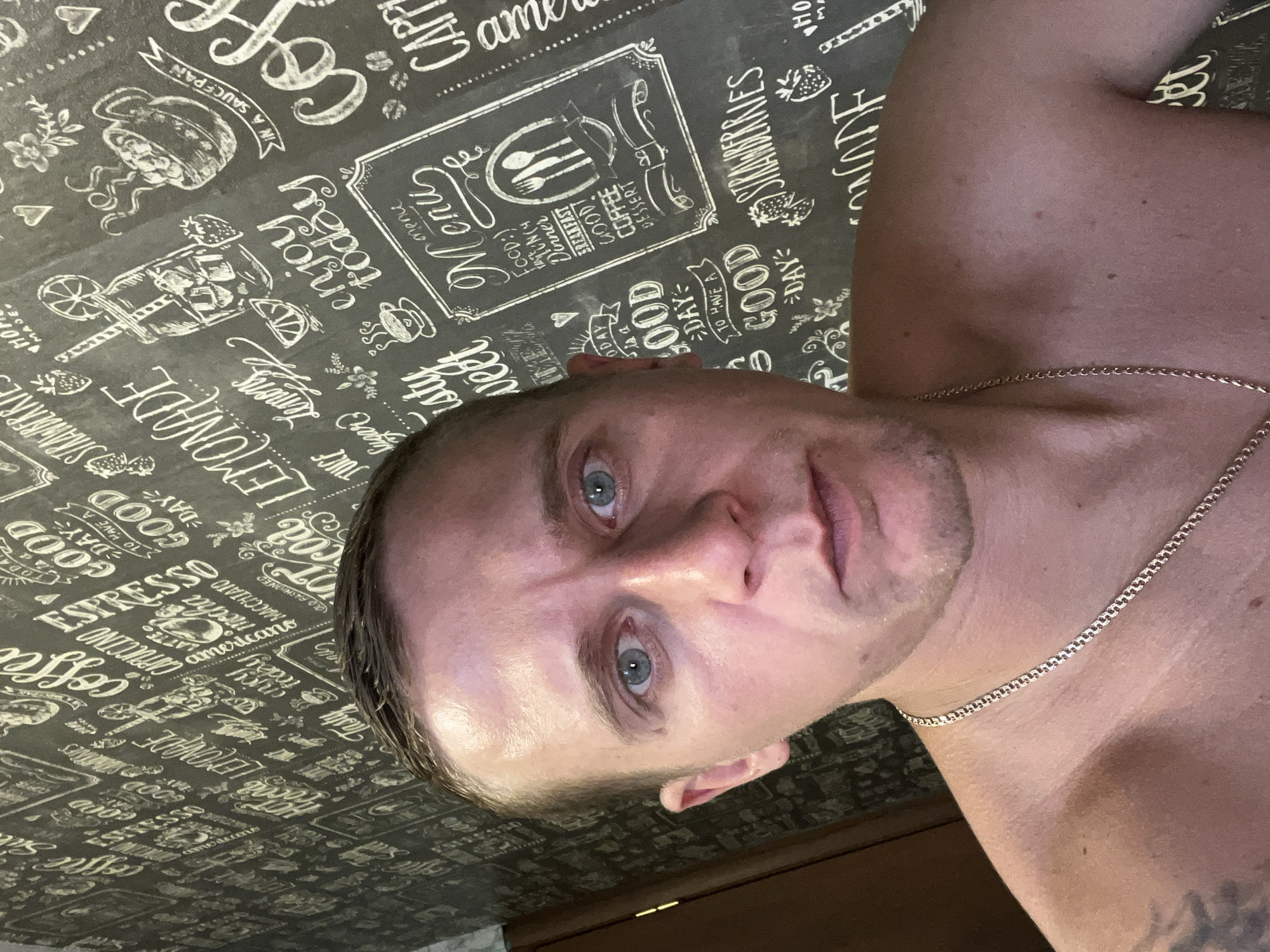 The image is a selfie taken by a shirtless man with short brown hair and blue eyes. He is wearing a silver chain necklace. The background is a wall with a chalkboard-style pattern featuring various coffee-related drawings and text, including "GOOD DAY," "LEMONADE," and "COFFEE." The setting appears to be indoors. No specific location in Minsk, Belarus, can be identified from the image.