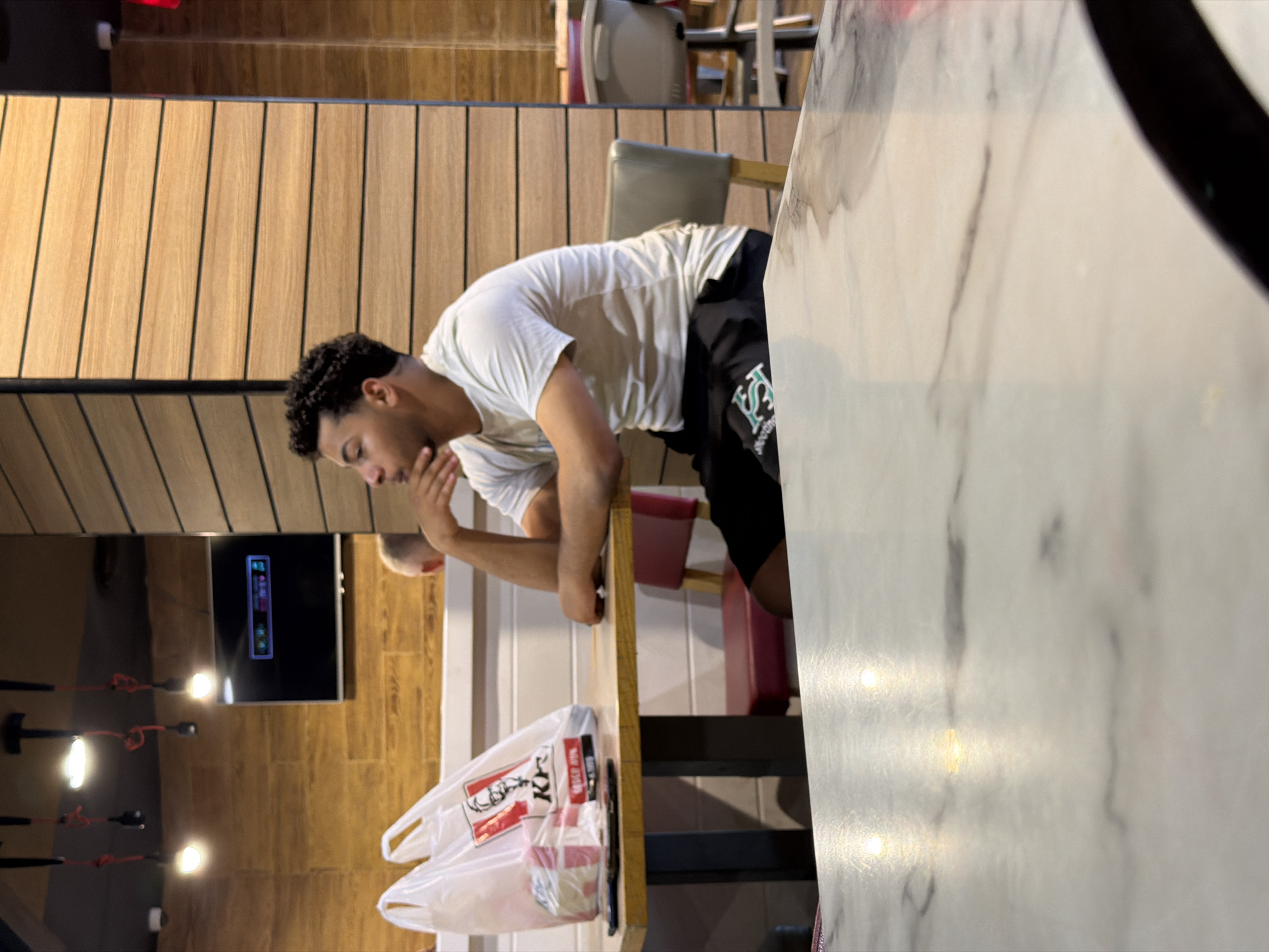 A young man of Middle Eastern descent is seated at a table in what appears to be a fast-food restaurant in Sharm El Sheikh, Egypt. He is wearing a light-colored t-shirt and black shorts with a green logo. His left arm is resting on the table, and he is looking to his right, with his chin resting on his hand, in a pensive pose. On the table in front of him are two white plastic bags from KFC, one of which contains food items. The table itself is wooden, and he is seated on a grey chair. In the background, a television screen displays a colorful graphic. The wall behind the man is paneled with horizontal wooden slats. The overall environment suggests a casual dining setting.