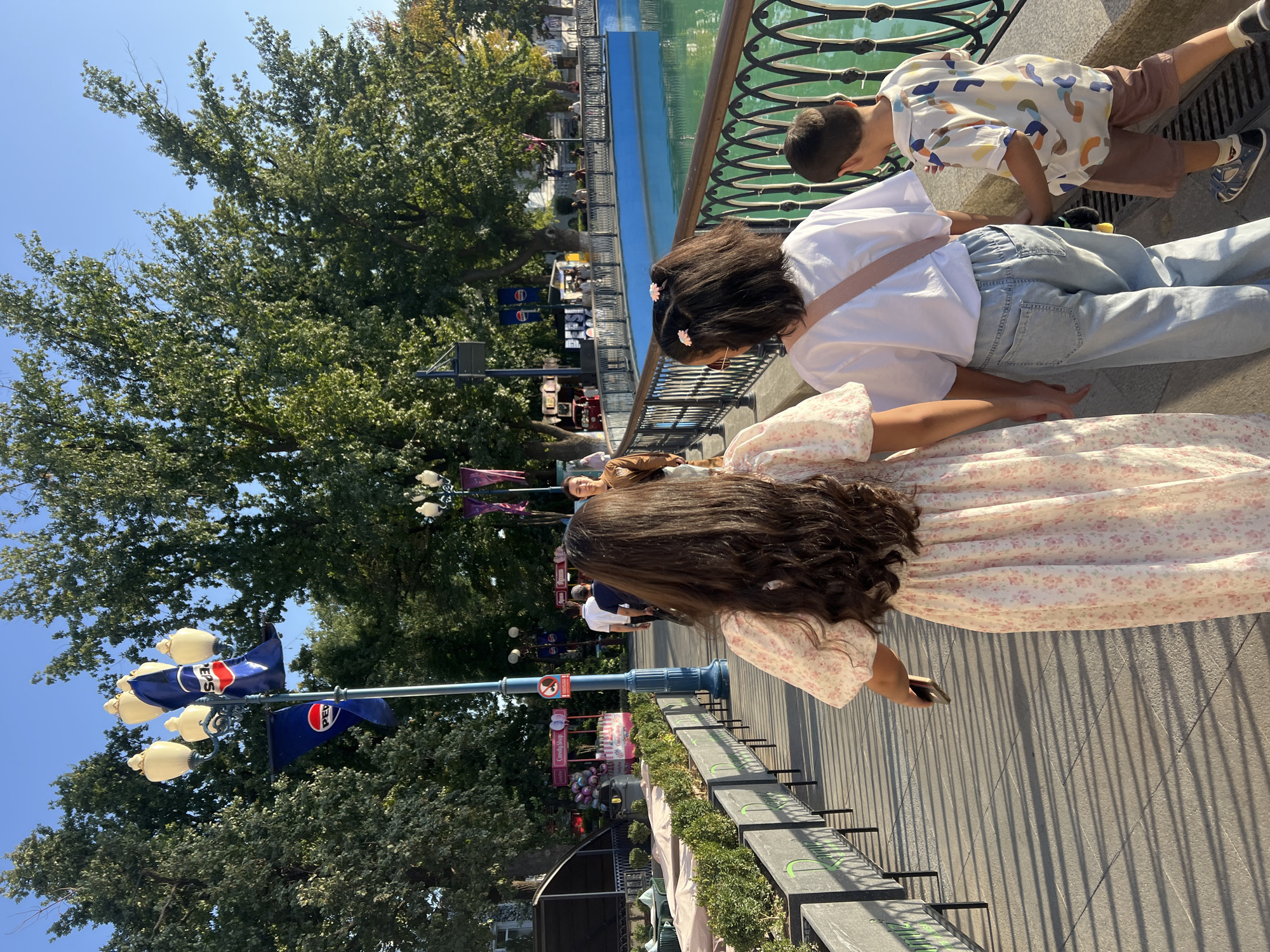 A group of three children and one adult are walking away from the camera on a paved pathway in an outdoor amusement park or recreation area. The children consist of two girls and one boy. The girl in the foreground on the left is wearing a long, floral-print dress and has long, dark hair. Beside her is another girl wearing a white t-shirt and light-wash jeans, with her hair in two pigtails. The boy on the right is wearing a colorful, patterned shirt and brown shorts. An adult is partially visible behind the children, walking further down the path. The pathway is lined with planters and a decorative metal railing overlooks a bright blue body of water. In the background, there are trees, lampposts with Pepsi flags, and what appear to be food stalls or small shops. The sky is clear and blue, suggesting a sunny day. The scene is illuminated by natural sunlight, casting distinct shadows from the railing onto the pathway.