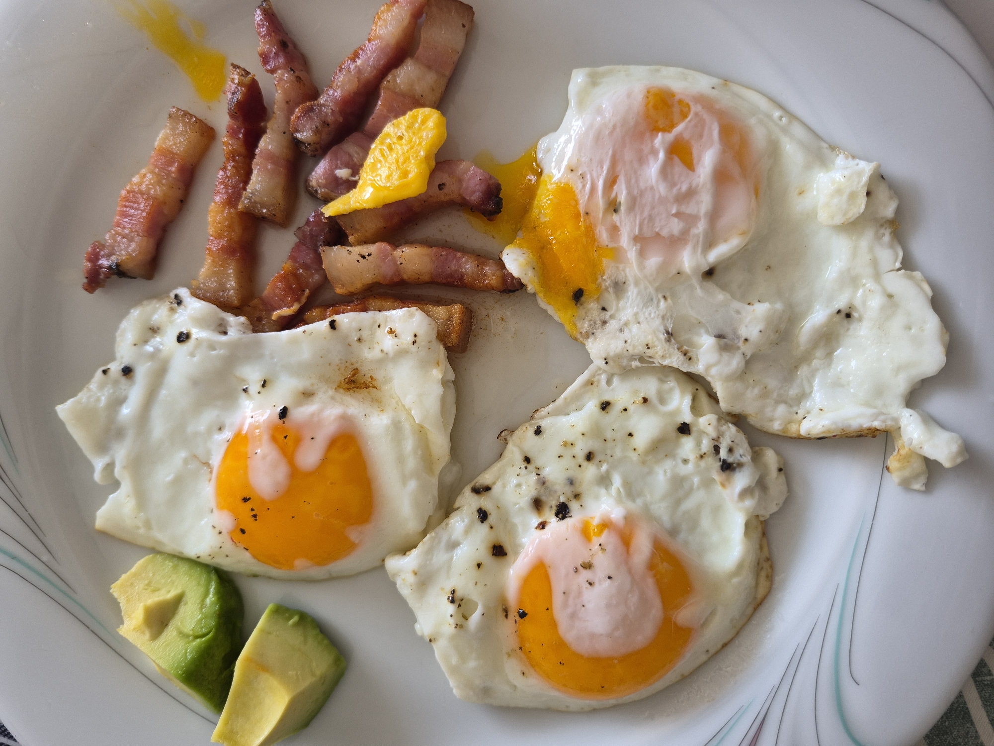 A plate of breakfast food is shown from a top-down perspective in an indoor setting. The plate contains three fried eggs, several strips of bacon, and two pieces of avocado. The fried eggs have sunny-side-up yolks and are seasoned with black pepper. The bacon strips are crispy and some have rendered fat. The avocado pieces are diced and are bright green on the outside and yellowish-green on the inside. The plate has a decorative rim with thin lines. The background is out of focus but appears to be a tablecloth.