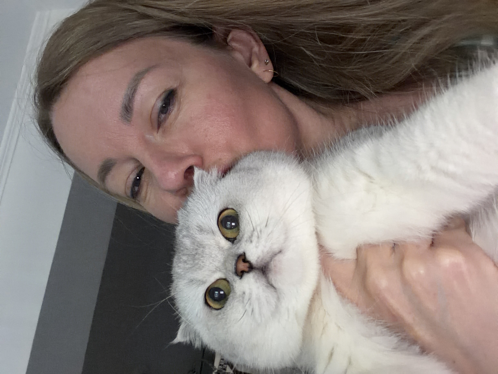 A woman is holding a fluffy white and grey cat to her face, appearing to kiss it. The woman has fair skin, long blonde hair, and is wearing a small hoop earring in her left ear. The cat has large, round, yellow eyes and a small pink nose. The background is out of focus, but appears to be an indoor setting with neutral colored walls and ceiling. There is no indication of the image being taken in Rome, Italy.