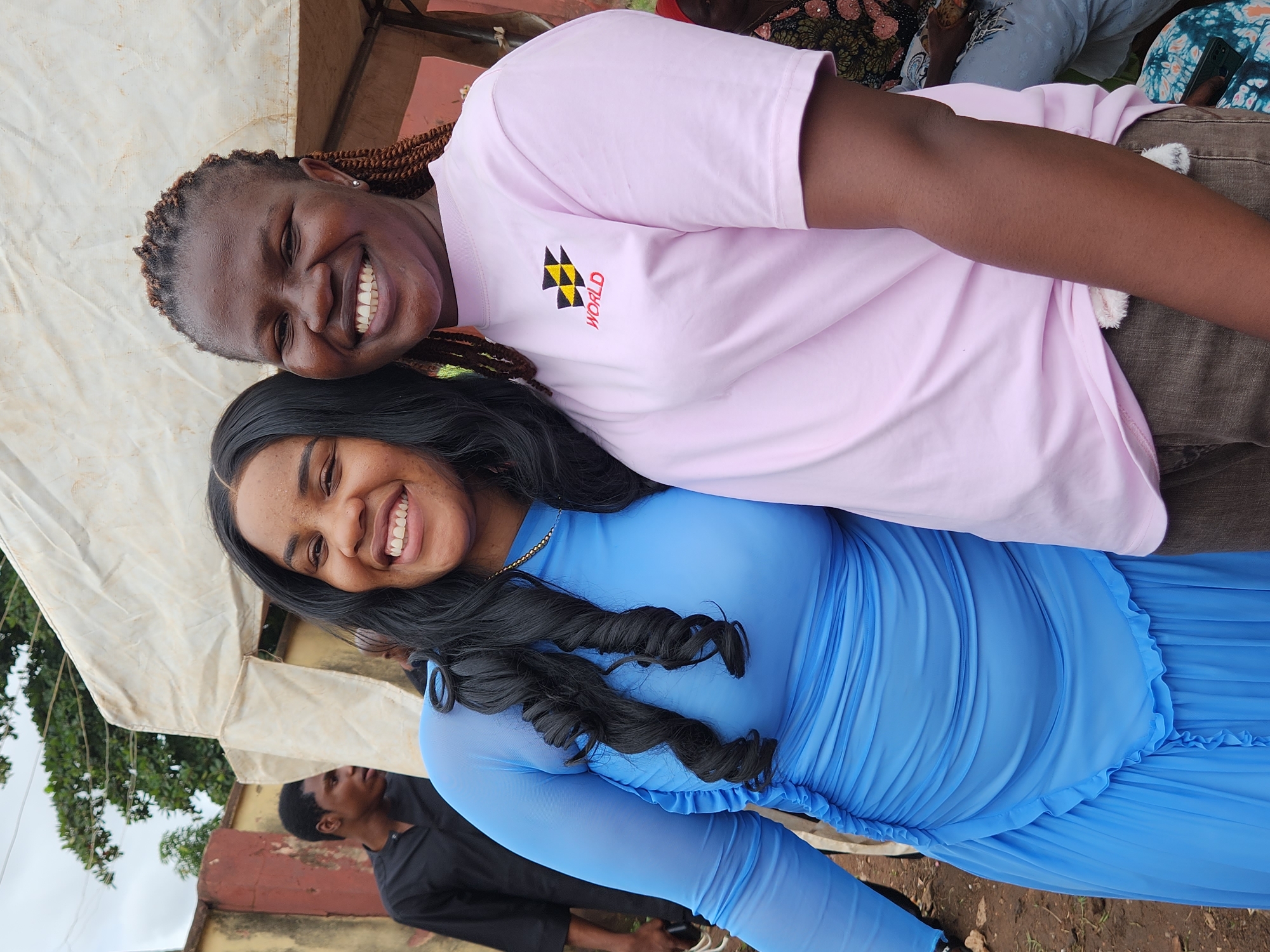 In Sabon Tasha, Nigeria, two Black women are captured in a candid outdoor portrait, smiling directly at the camera. The woman on the left is wearing a long, flowing, bright blue dress with a ruched bodice and bell sleeves. Her dark, curly hair cascades over her shoulders. The woman on the right is wearing a light pink t-shirt with a small emblem and the word "WORLD" printed on it. Her hair is styled in braids, and she is wearing a silver stud earring in her visible ear. Both women appear to be in good spirits.
Behind them, a makeshift canopy made of white fabric provides shade. A man in a black shirt is visible in the background, to the left, his back to the camera. The environment appears to be unpaved, with dirt and scattered debris on the ground. A partial view of a red-brick building is visible to the far left, behind the man in black. The overall scene suggests a casual gathering or event taking place in a semi-urban or rural setting.