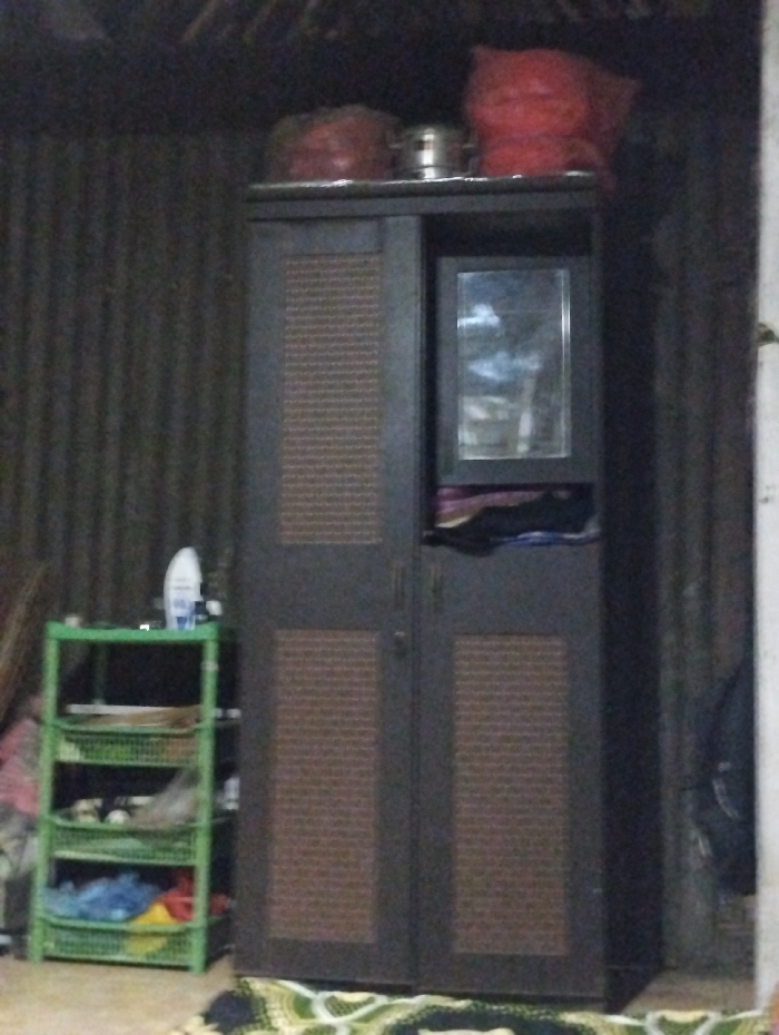 The image captures an indoor scene in Kecamatan Arjasa, Indonesia, characterized by dim lighting. The central focus is a dark brown, two-door wardrobe or cabinet. Its doors feature rectangular panels with a lighter brown, textured or slatted design. An integrated mirror is visible on the upper right door of the cabinet, reflecting a blurry, light-colored area.
On top of the wardrobe, there are several items: two reddish fabric bundles or bags, and a metallic pot positioned between them. To the left of the wardrobe stands a light green, four-tiered plastic shelving unit. The top shelf holds a white bottle, while the lower shelves contain various household items, including what appear to be clothes or other articles stored in baskets.
The background wall is constructed from dark, corrugated metal sheets, suggesting a simple or utilitarian dwelling. A colorful, patterned rug or mat lies on the floor in the foreground. No people are present, and no specific activity is depicted. The subdued lighting indicates the photograph was taken indoors with limited light, possibly in the evening or in an unlit room. No visible text is clearly legible.