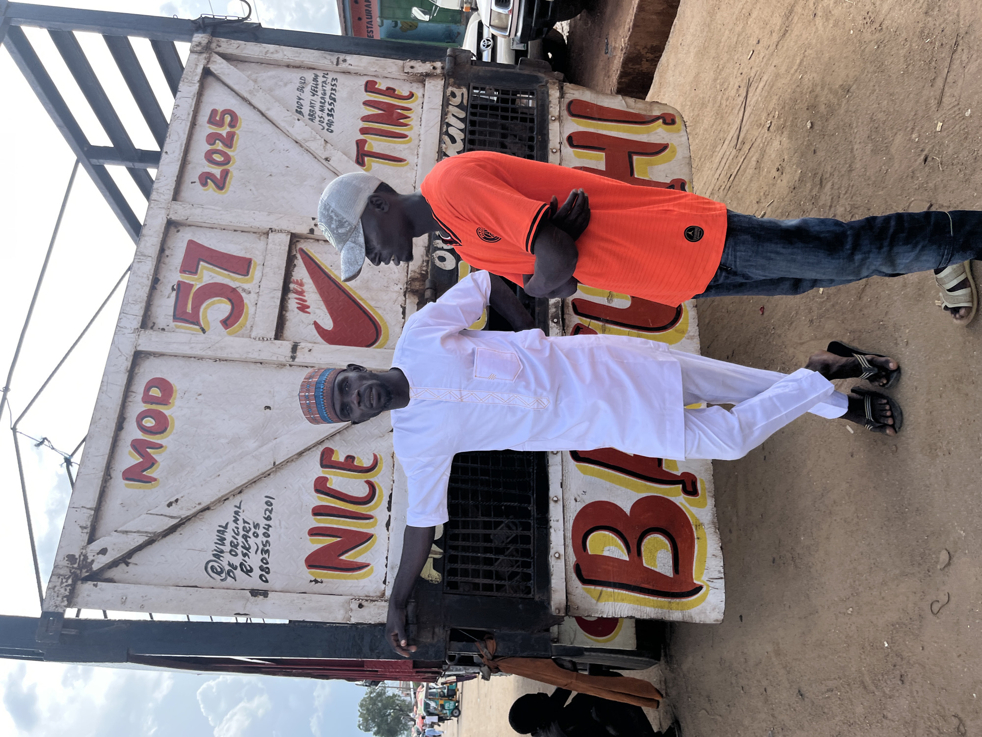 Two men stand in front of a weathered white truck in Jos, Nigeria. The truck's side is adorned with hand-painted lettering and graphics, including the words "MOD," "57," "2025," "NICE," and "BAGI." A Nike logo is also visible. The man on the left wears a white traditional outfit and a patterned cap, while the man on the right is in an orange t-shirt, a grey baseball cap, and blue jeans. In the background, other trucks, a tricycle, and people are faintly visible, suggesting a marketplace or transportation hub. The ground is dusty and uneven. The man on the left has his left arm resting on the truck. The man on the right has his arms crossed.
