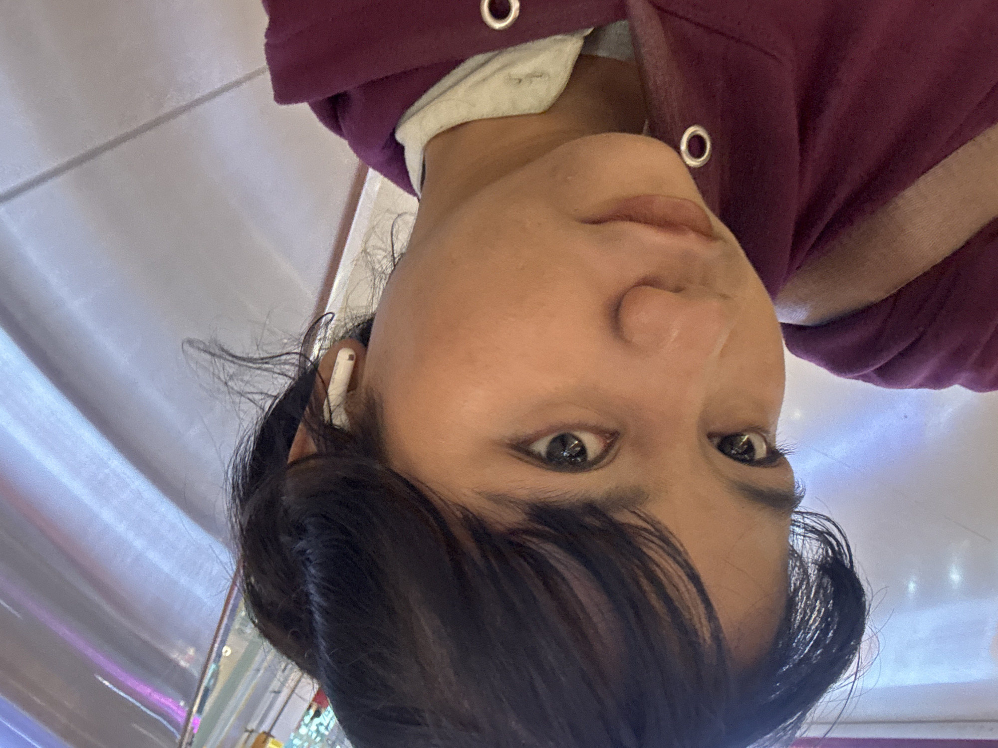 A close-up, angled, selfie-style shot shows the face of a young person with dark hair, wearing a burgundy hooded sweatshirt and a white t-shirt. The person has dark eyes and is wearing white wireless earbuds. The background appears to be an indoor setting with a reflective, curved ceiling or wall with horizontal lines and hints of purple and blue lighting. The image was taken in the city of North Jakarta, Indonesia.