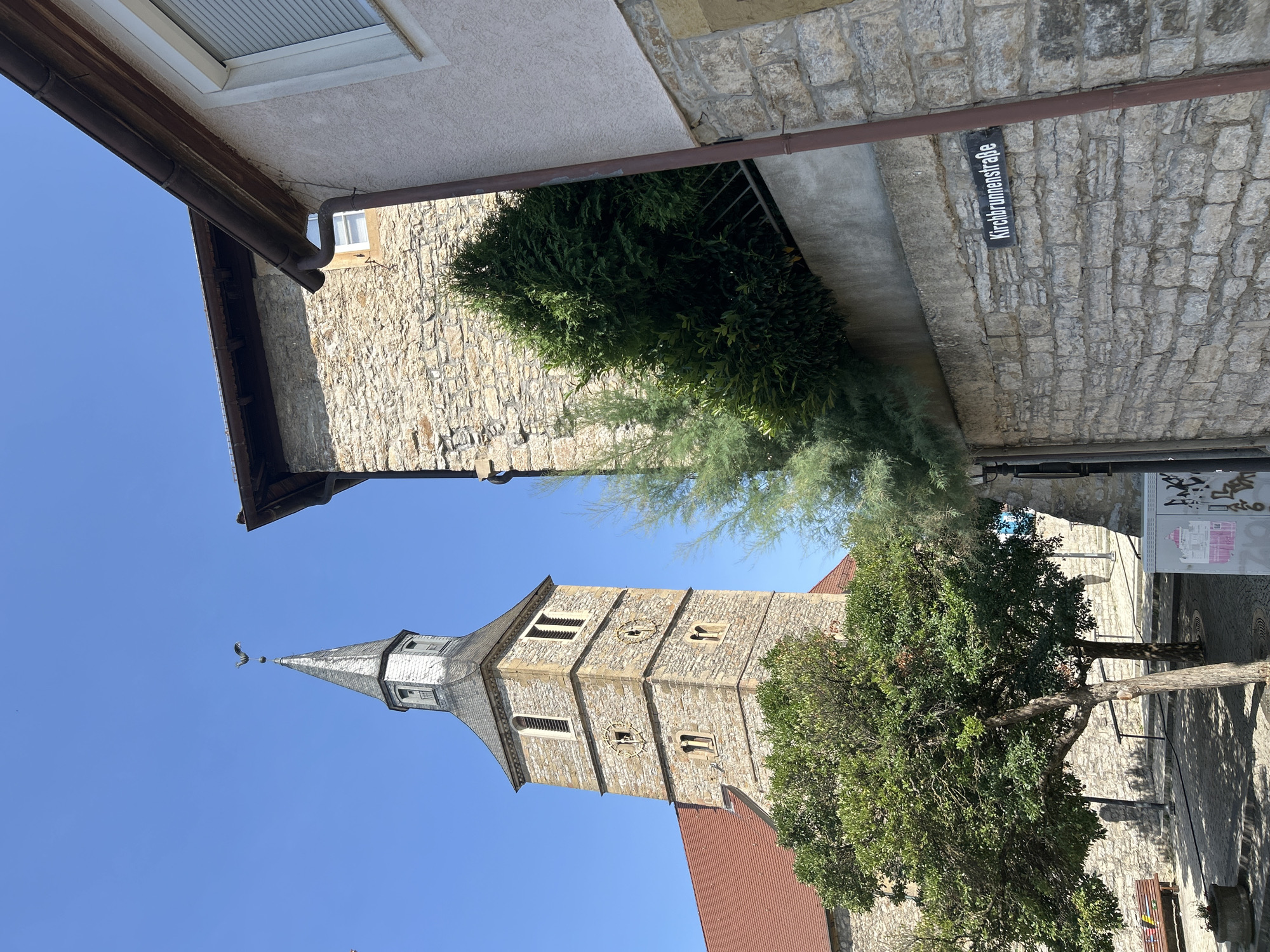 The image captures a street scene in Bad Friedrichshall, Germany, featuring a prominent church tower with a weather vane. The tower is constructed of stone blocks and has a steep, grey roof. Adjacent to the tower, a modern building with a light-colored facade and a brown gutter system is visible. The street itself is paved with cobblestones. Green trees and bushes add to the environment. A street sign reading "Kirchbrunnenstraße" is affixed to a stone wall. The sky is clear and blue.