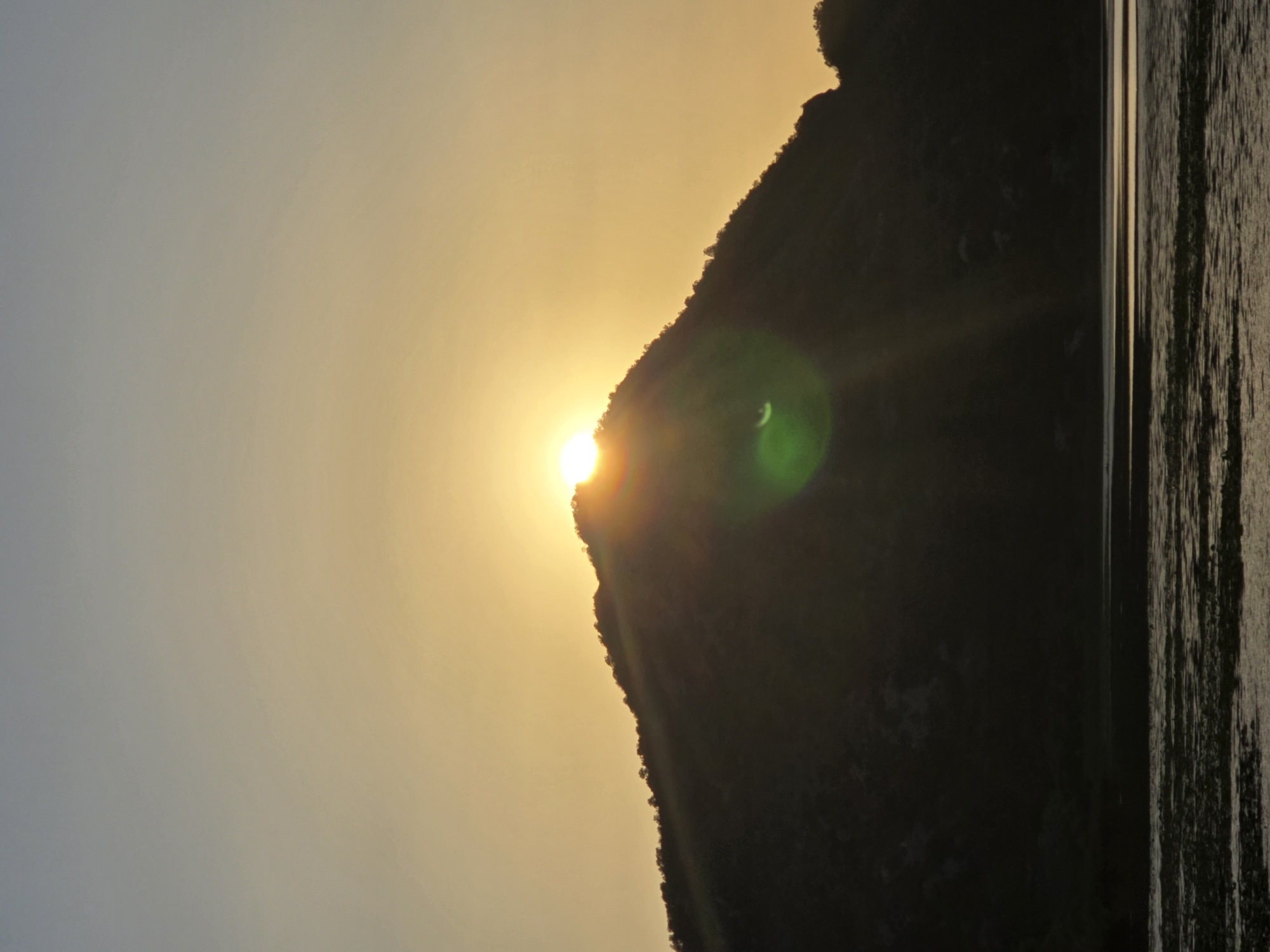 The image shows a sunset over a mountainous landscape with a body of water in the foreground. The sun is a bright orb peeking over the crest of a dark, silhouetted mountain, casting a warm, golden glow across the sky. The sky transitions from a soft yellow near the horizon to a muted gray at the top of the frame. A lens flare, appearing as a green orb, is visible to the right of the sun. The mountain itself is covered in dark vegetation, its silhouette sharply defined against the bright sky. The body of water in the foreground appears calm, with ripples reflecting the subdued light. Patches of aquatic vegetation are visible on the surface of the water. The scene is tranquil and evokes a sense of natural beauty. The location is identified as Seoca, Montenegro.