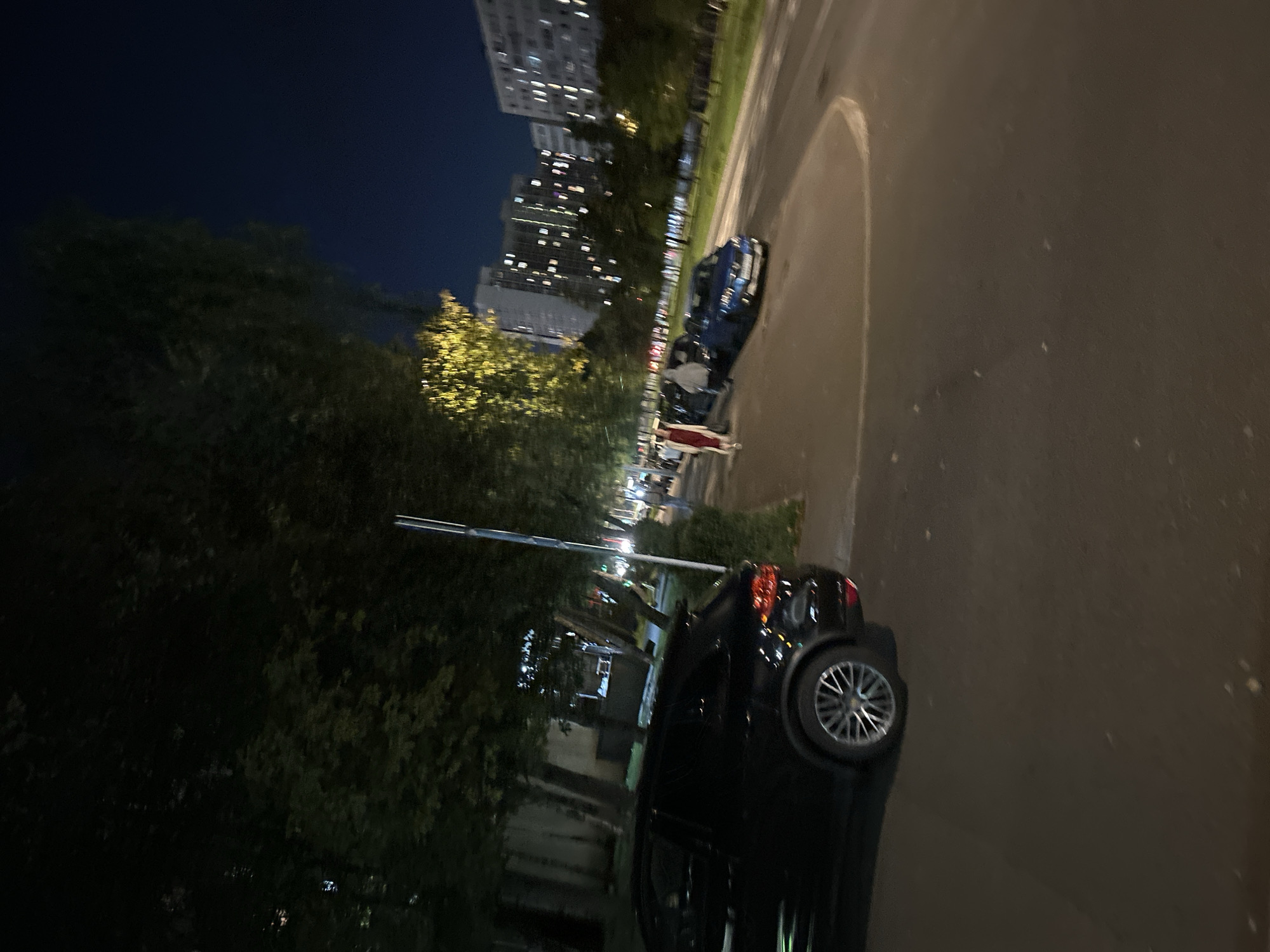 The image shows a street scene at night, possibly in Moscow, Russia. In the foreground, a black SUV is parked on the left side of the road. The vehicle is dark and the reflections on its surface are minimal. Beyond the SUV, a sidewalk runs alongside the road, lined with trees. Several people are visible walking on the sidewalk, some of them appearing to be in groups. Further down the street, a blue car is parked, and more people are near it. In the background, tall apartment buildings with illuminated windows dominate the skyline, suggesting an urban environment. The sky is dark, indicating it is nighttime. Streetlights and lights from the buildings cast a dim glow on the scene. The overall impression is of a residential or commercial street in a city after dark.