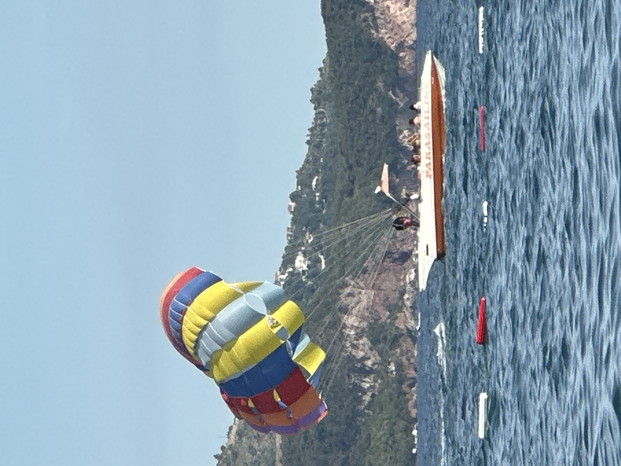 In the city of Boreti, Montenegro, a brightly colored parachute, featuring red, orange, yellow, blue, and purple panels, is suspended in the air. It is attached by multiple ropes to a white speedboat with an orange stripe, which is navigating the choppy blue water. Several individuals are visible on the boat, appearing to be passengers or crew members. The word "PARASAILING" is visible on the side of the boat. In the background, a green, tree-covered mountainside with scattered white buildings rises from the coastline. The sky above is a clear, light blue. In the foreground, two rectangular buoys, one white and one red, float on the surface of the water.