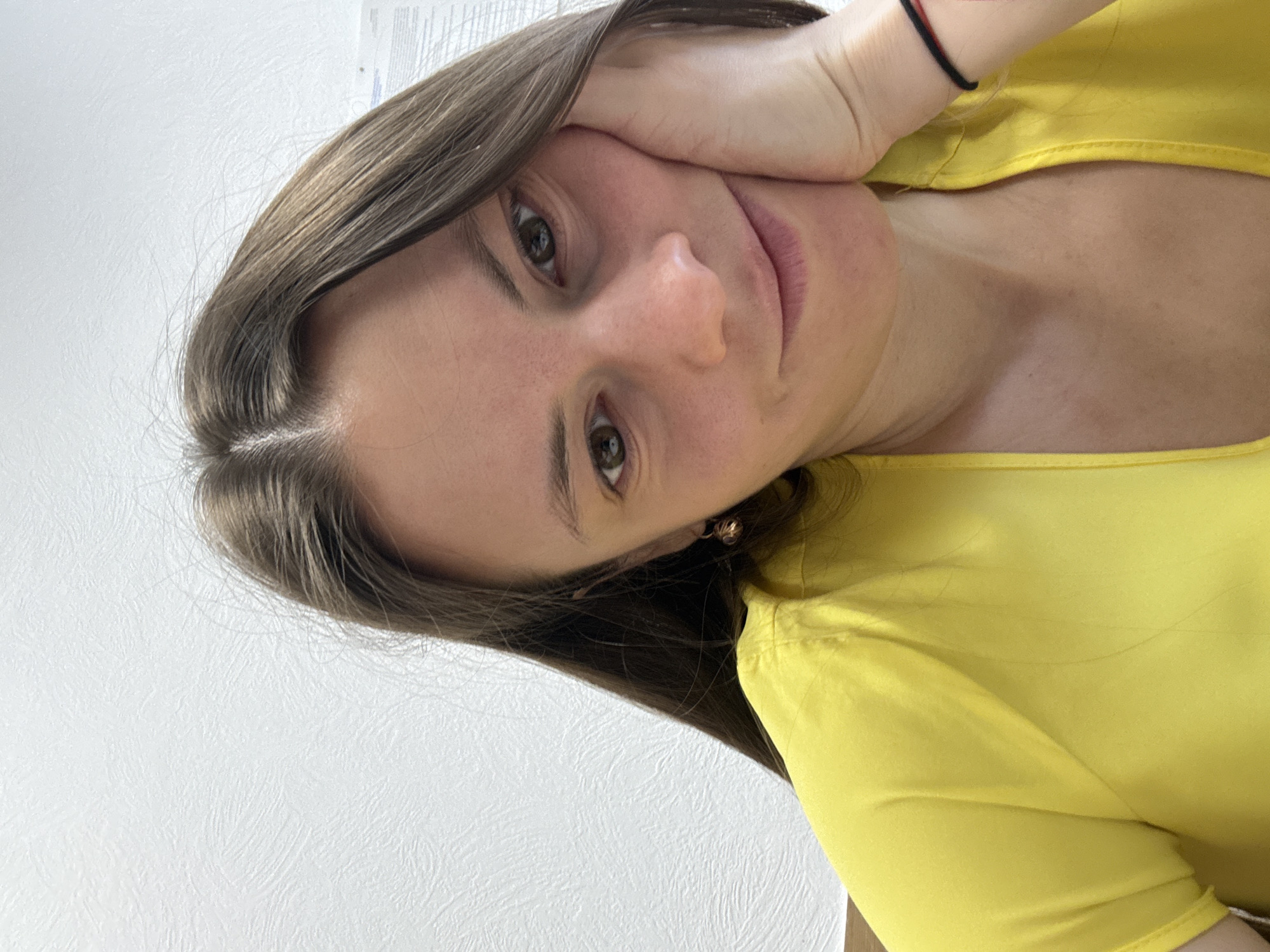 A young woman with brown hair and fair skin is posing for a selfie indoors. She is wearing a yellow top and has her left hand resting on her face. She is wearing a black bracelet on her left wrist and gold earrings in her left ear. The background is a plain white wall. The city of Chaikovsky, Russia, is not visible in the image.