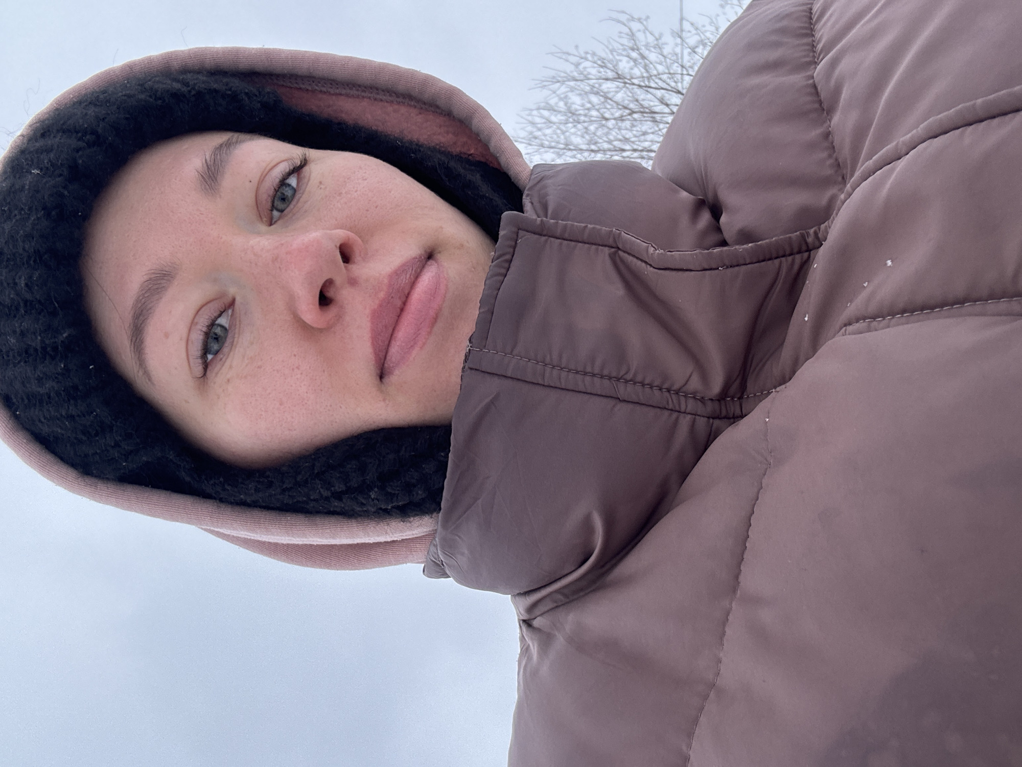 A close-up, low-angle shot shows a woman wearing a puffy, taupe-colored winter coat with a hood. The hood is lined with black fleece and has a pinkish-brown outer layer. The woman has fair skin, freckles, light eyes, and full lips. She is looking downward toward the camera with a slight smile. The background consists of a pale sky and bare branches of a tree. Based on the sky color and the type of clothing worn, the location appears to be cold, possibly winter. The location is in the city of Ферма, Russia.