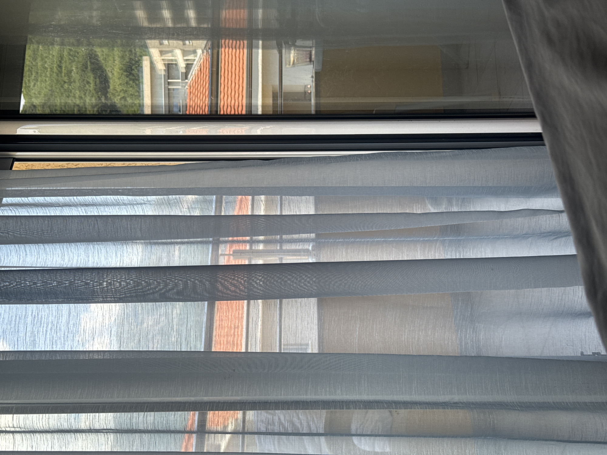 The image shows a view through a sheer curtain in Budva, Montenegro. The curtain is light blue and hangs in vertical folds, obscuring a portion of the scene outside. Through the curtain and a glass sliding door, the viewer can see a building with an orange-tiled roof. Further in the distance, a lush green hillside rises. Reflections are visible on the glass, showing parts of the interior and the exterior environment. A small glass with some liquid is on a ledge visible through the glass. The foreground shows a gray, textured surface, possibly a bed or furniture.