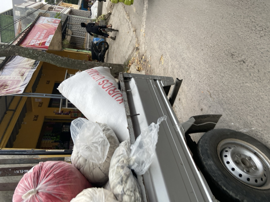 The image shows the back of a silver pickup truck parked on a street in Margaasih, Indonesia, at the coordinates -6.955, 107.55. The truck is loaded with large white bags, a red bag, and white plastic bags. The bags are stacked on the truck bed. The street is paved with asphalt. A man is walking away from the camera on the sidewalk, along a wall with a sign that reads "Makmur Barokah Daya". A tree is in the foreground, and a yellow building is visible in the background. The pickup truck is facing towards the right, and the back tire is visible. The back tire is partially obscured by a large white bag marked with the words "KURIOS UTAMA".