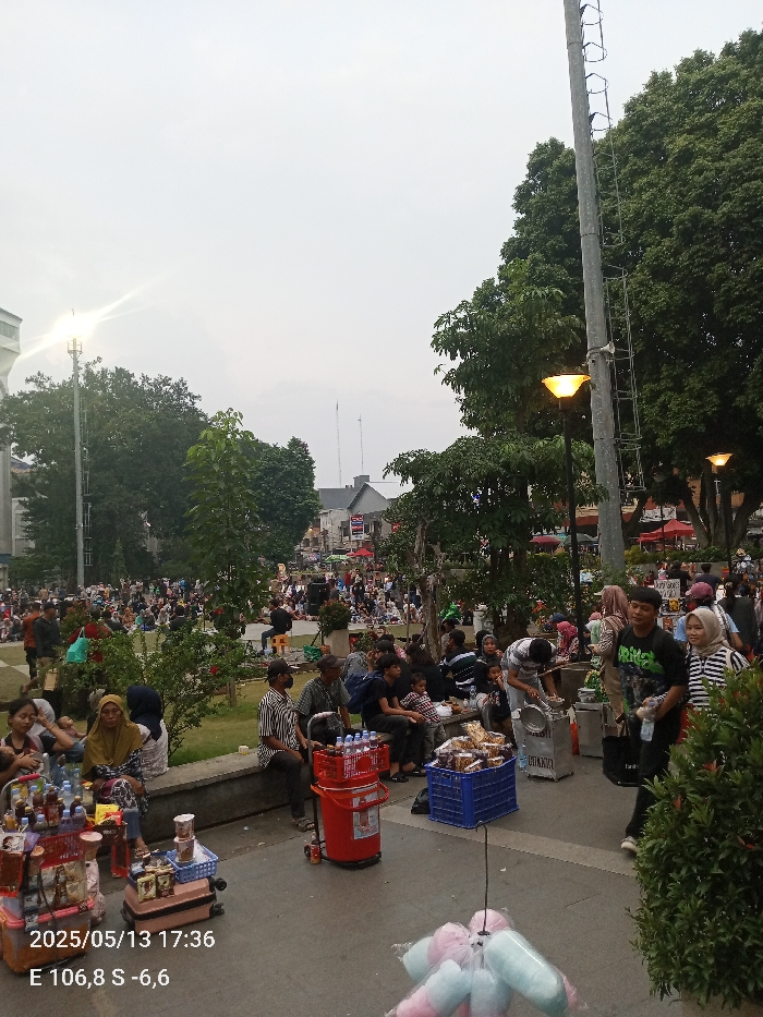 The image shows a street scene in Bogor, Indonesia. The coordinates are: latitude -6.595, longitude 106.792. The scene is a busy street with many people walking and sitting on benches. There is a street vendor selling drinks and snacks. There are many trees and a street lamp in the background. The time is 5:36 PM on May 13, 2023. A street vendor is selling drinks and snacks from a cart. There are people walking past, and many people are sitting and talking on benches. There are a few other street vendors in the background, and many green trees line the street.