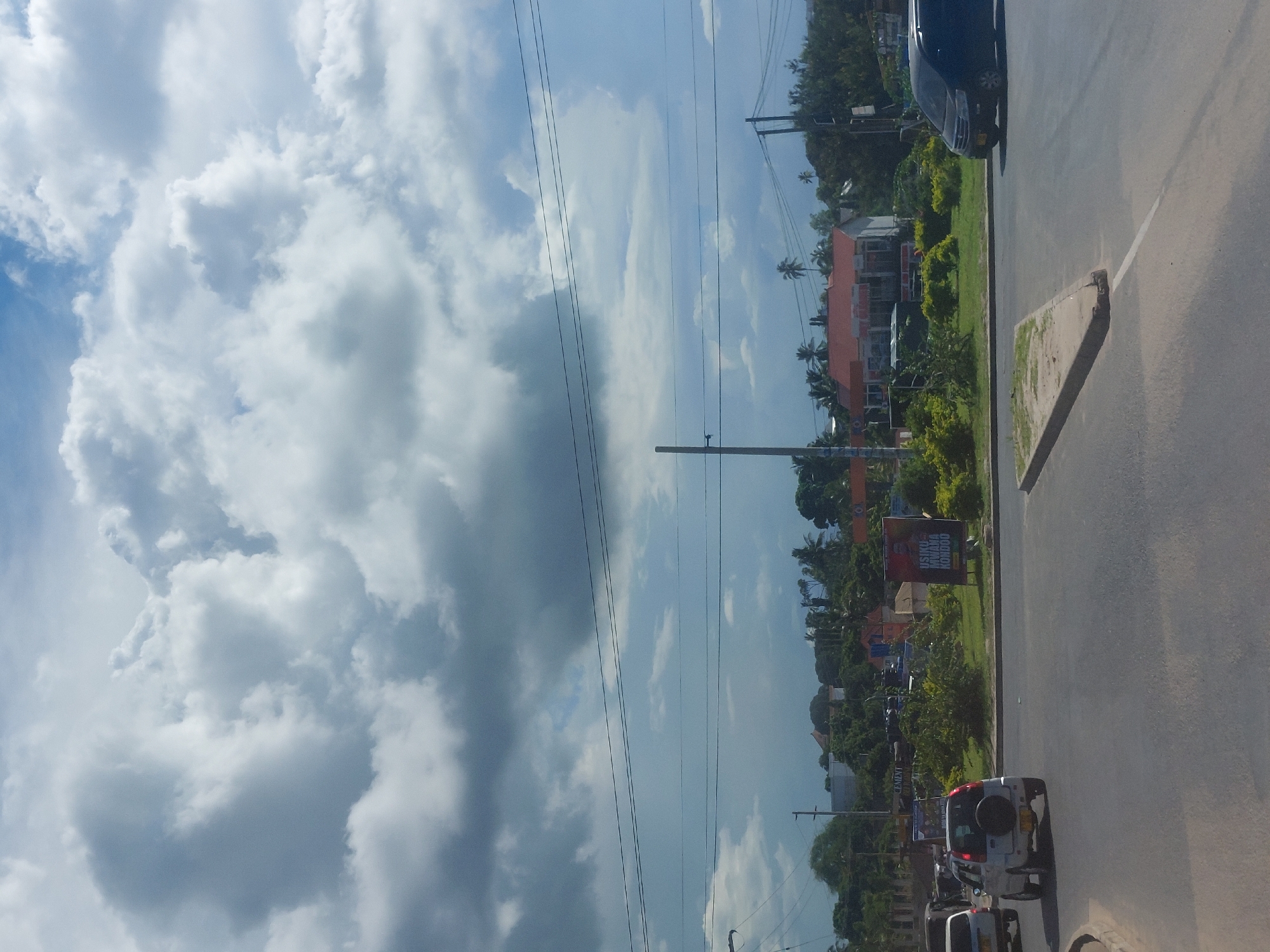 The image presents a daytime exterior view of a multi-lane asphalt road in Dar es-Salaam, Tanzania, beneath a partially cloudy sky with large cumulus formations and visible blue patches. In the foreground, a triangular concrete median with a white painted line separates road lanes. A light-colored compact SUV with a rear-mounted spare tire is observed from its rear-left quarter, moving leftward. On the right side of the road, a dark-colored van is positioned, facing right. No individuals are visible.
The central median is vegetated with green bushes and small trees. Two billboards are located on this median: a prominent one on the right displays the Swahili text "USIKU MWAWA MONDOO," and a smaller, partially obscured sign on the left includes the word "CEMENT." Behind the median, several buildings are visible, notably a structure with a reddish-orange roof and white walls. Overhead power lines and multiple utility poles traverse the scene horizontally and vertically. The background consists of dense tropical foliage, including palm trees, interspersed with additional structures.