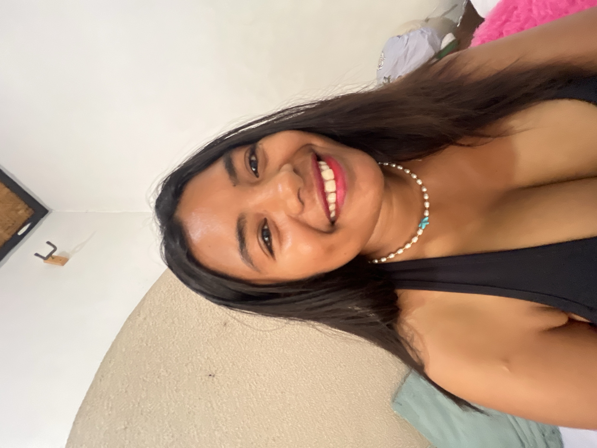 A young woman with long, dark hair smiles at the camera, wearing a black tank top and a pearl necklace with turquoise beads. She is indoors, with a textured wall behind her and a white wall to her right. The background suggests a bedroom or living space, possibly in Denpasar, Indonesia, given the lighting and tropical feel. There is a hint of greenery from a pillow on her left and a bright pink fuzzy object on her right. A simple wooden hook is mounted on the wall to the left of the frame. The overall atmosphere is casual and relaxed.