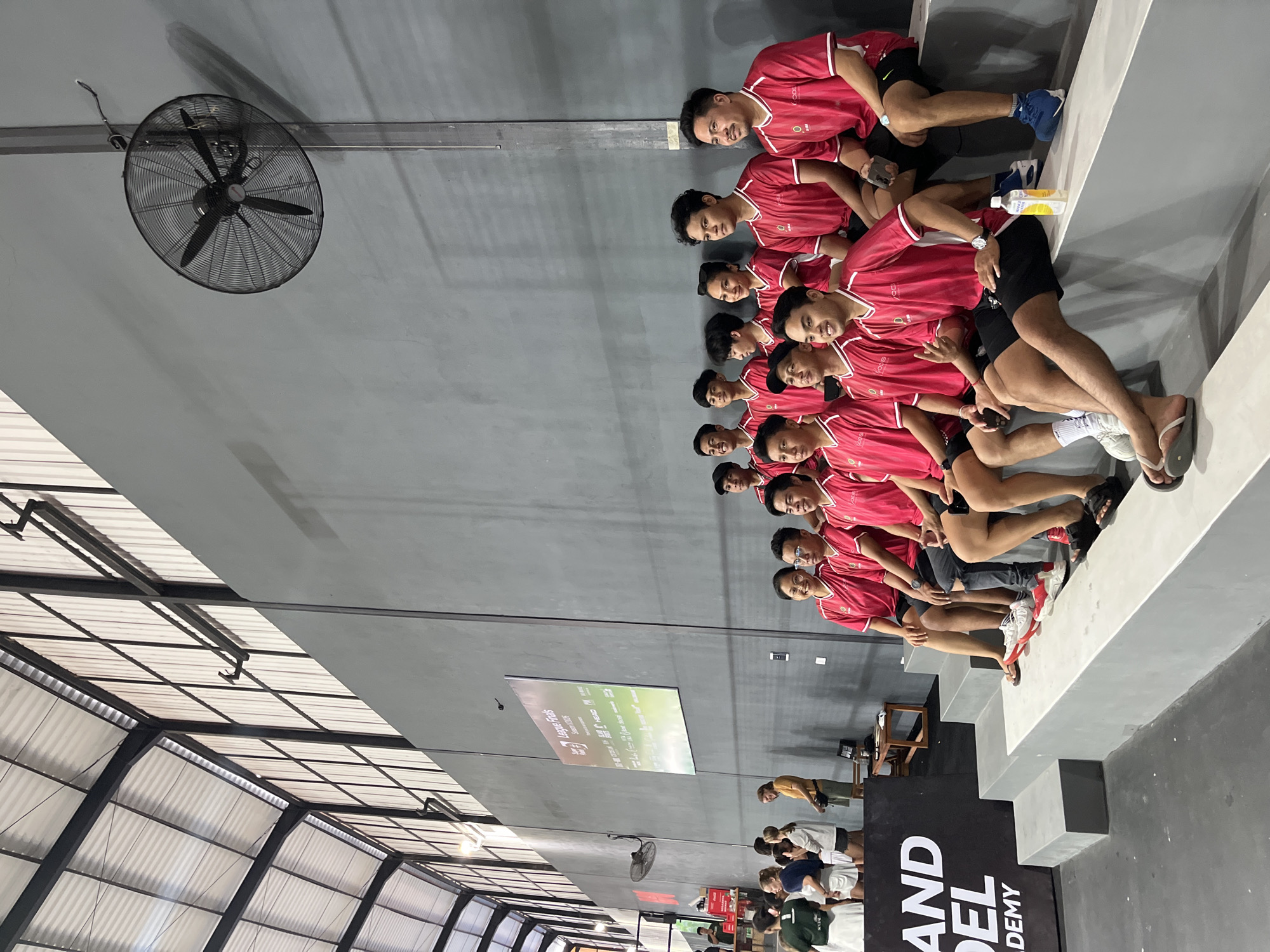 The image shows a group of people posing indoors. They appear to be in North Kuta, Indonesia, judging by the background. The scene consists of a group of at least twelve individuals, likely males, sitting on a concrete structure that resembles steps. They are wearing matching red shirts with white accents on the sleeves and neck, along with dark-colored shorts. The men seem to be posing for a group photograph. Behind them, there is a gray wall with a large screen displaying text in a foreign language. A large industrial fan is mounted on the ceiling. The location seems to be a semi-indoor space with a partially covered roof made of metal beams and translucent panels. In the lower portion of the image, there are more people standing and observing, near a stand marked "AND DEL ACADEMY."